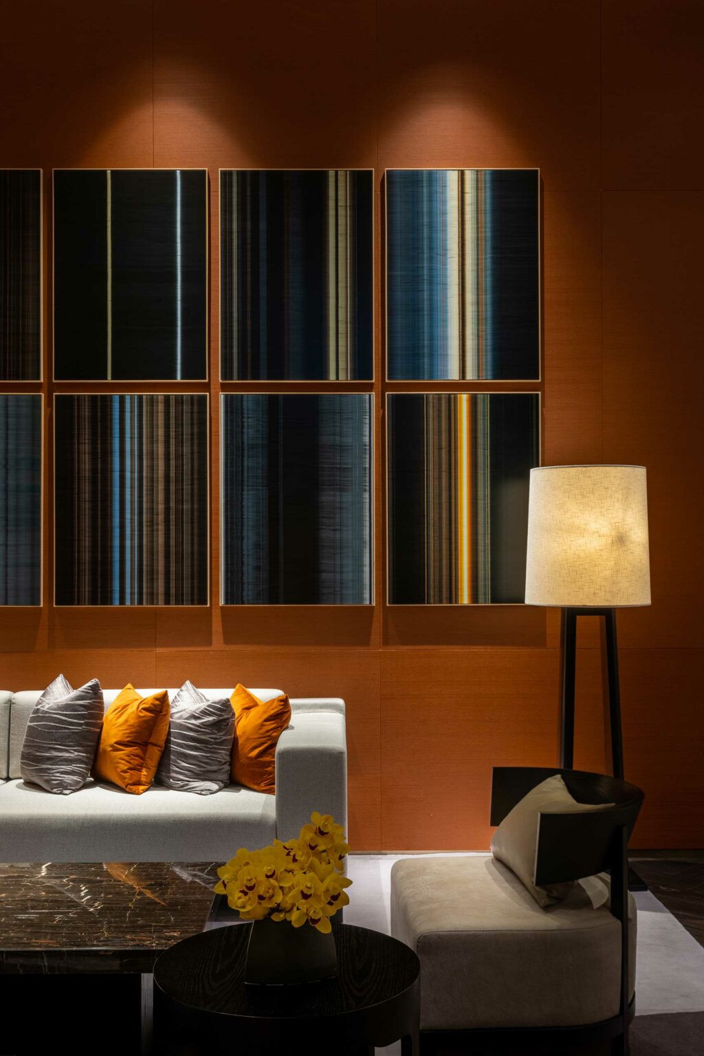 Contemporary hotel lounge with modern art panels featuring vertical stripes, a white sofa with orange and gray pillows, a floor lamp, and a round table with yellow flowers.