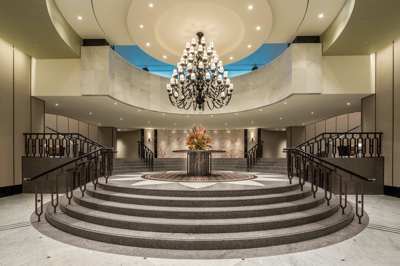 Elegant hotel ballroom lobby featuring a grand staircase leading to a large chandelier. Central flower arrangement adds a touch of warmth and sophistication.