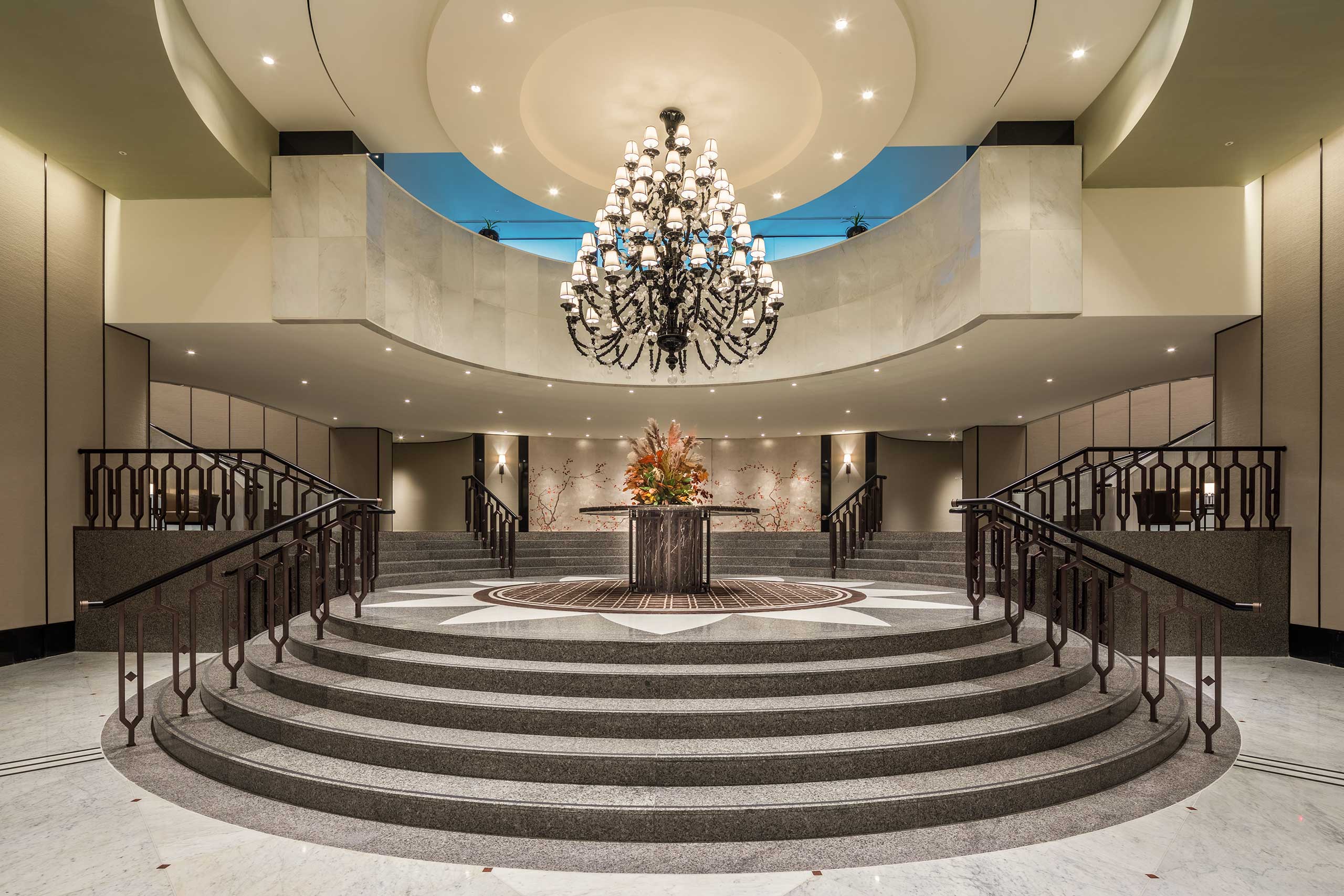 Elegant hotel ballroom lobby featuring a grand staircase leading to a large chandelier. Central flower arrangement adds a touch of warmth and sophistication.