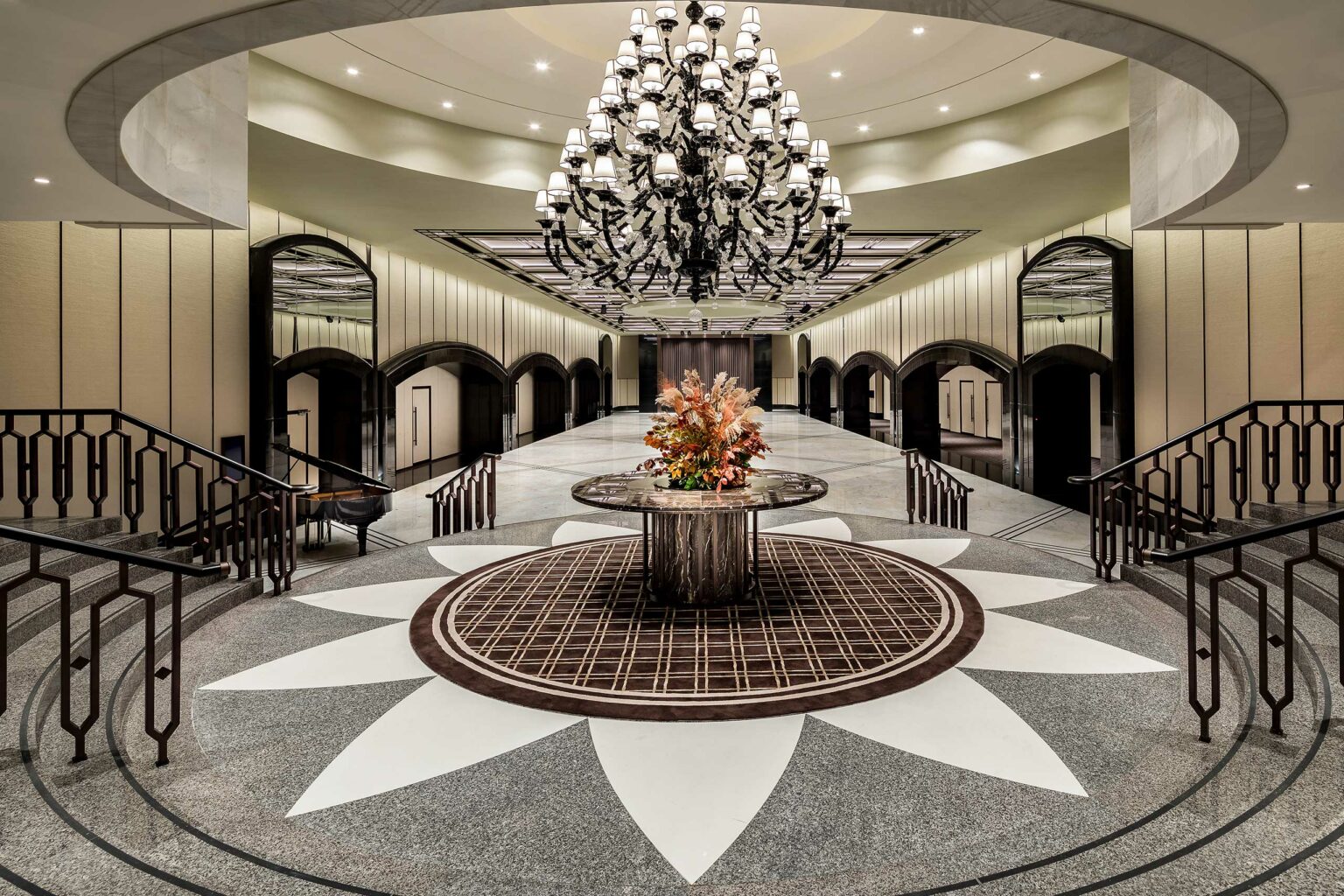 Elegant hotel ballroom lobby with a large floral arrangement on a round table, surrounded by symmetrical staircases. A grand chandelier hangs overhead, creating a luxurious atmosphere.