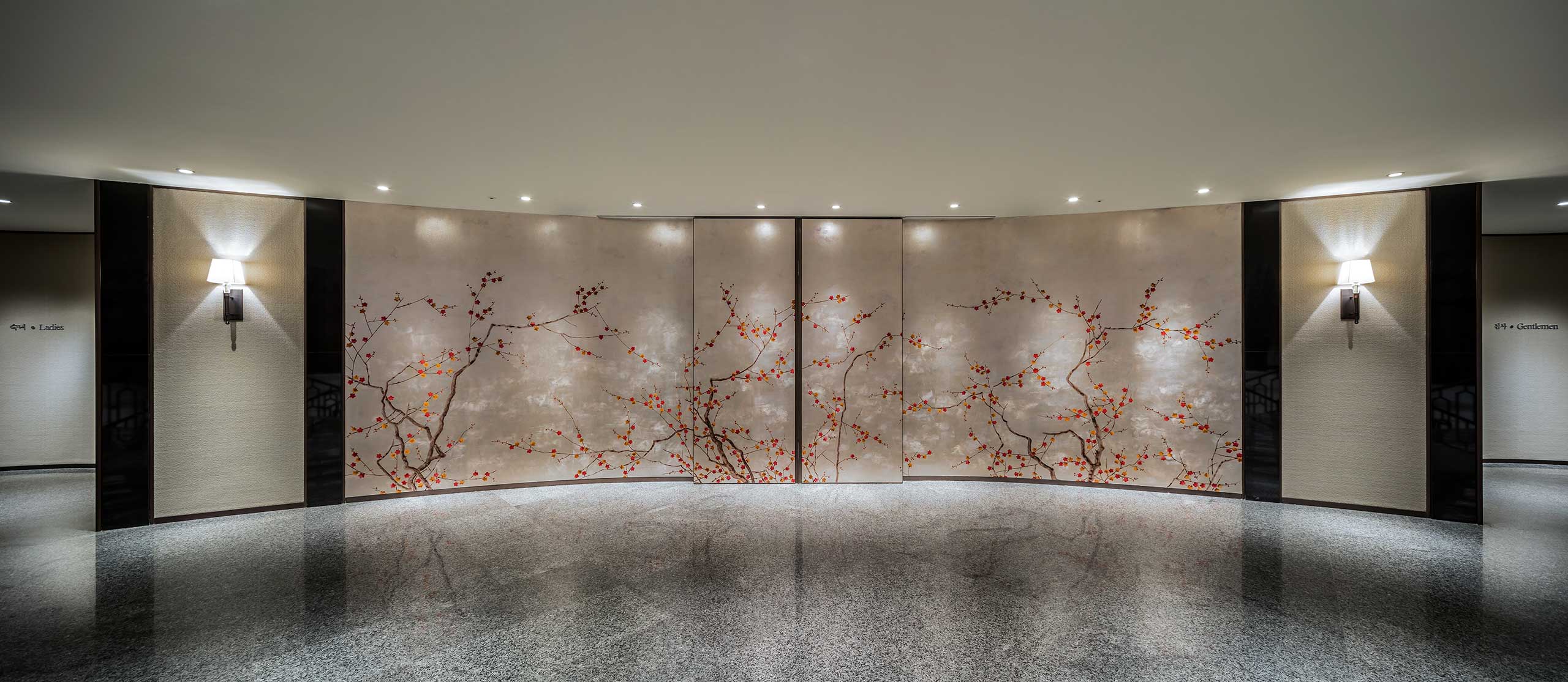 Hotel ballroom area featuring a curved wall with a floral mural depicting delicate branches and red flowers. Soft lighting from sconces creates a calm, elegant ambiance.