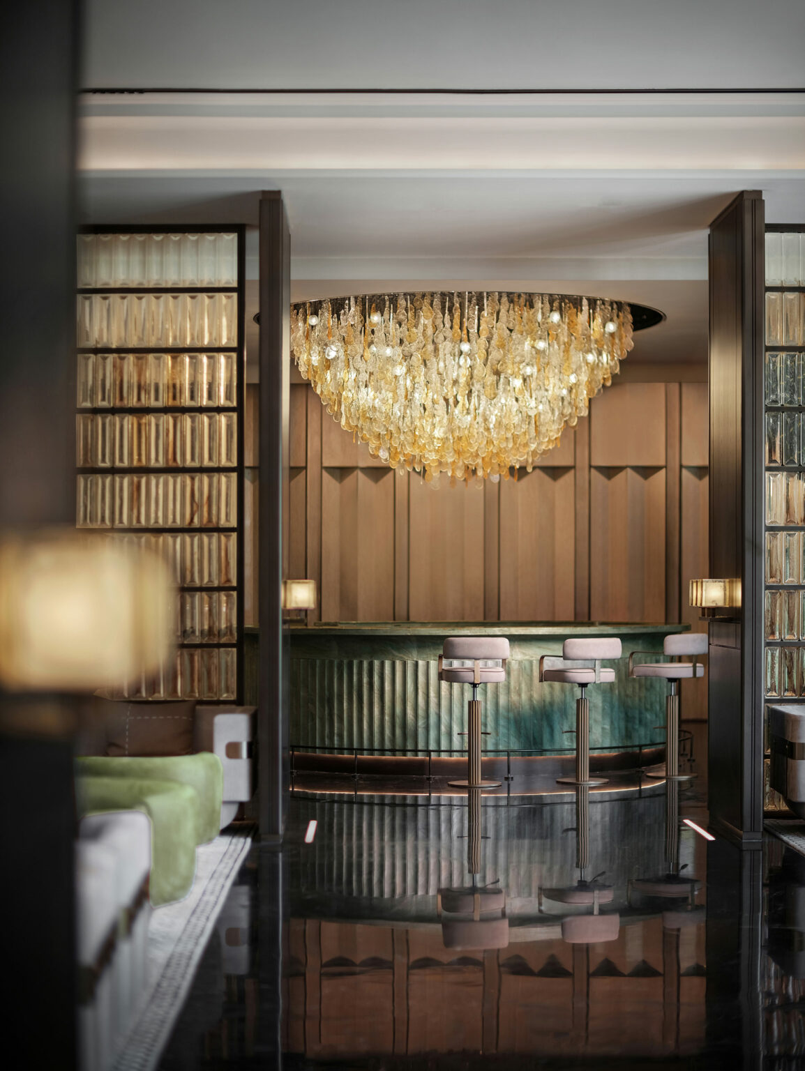 Luxurious bar with a large, ornate chandelier casting warm light over plush barstools and a curved, textured counter, creating an elegant atmosphere.