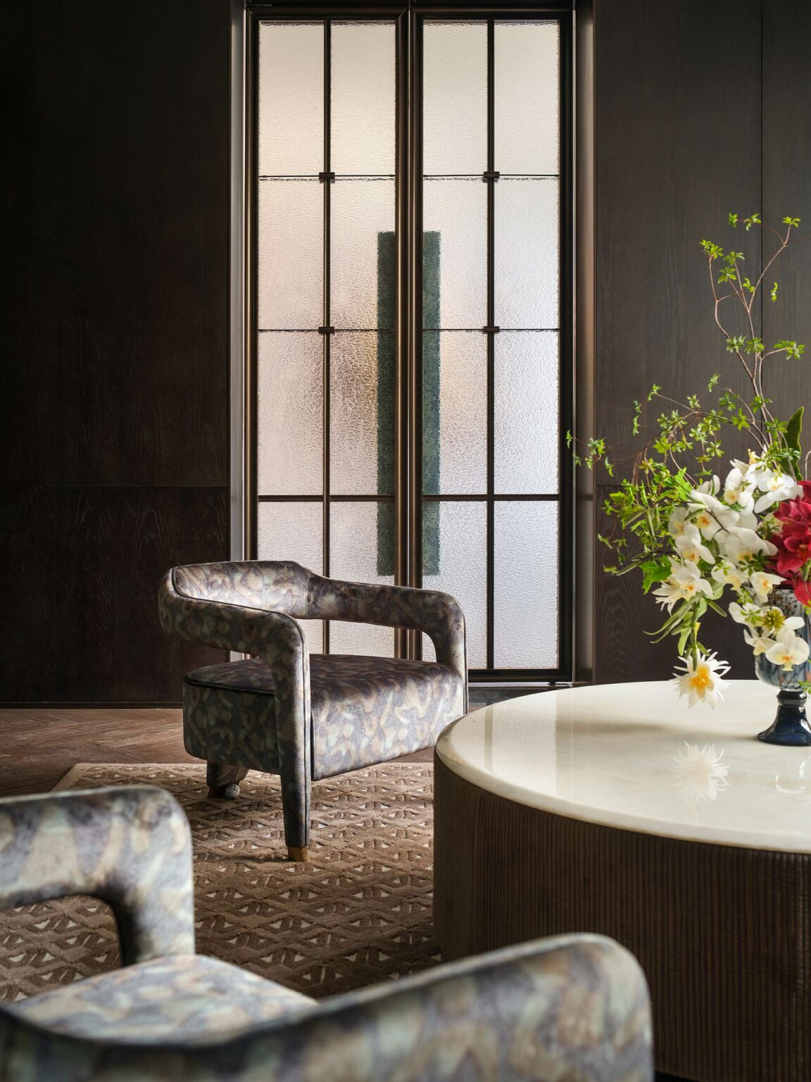 Chic living room with a frosted glass door, patterned armchair on a textured rug, and round table with a vibrant floral arrangement, creating a serene ambiance.