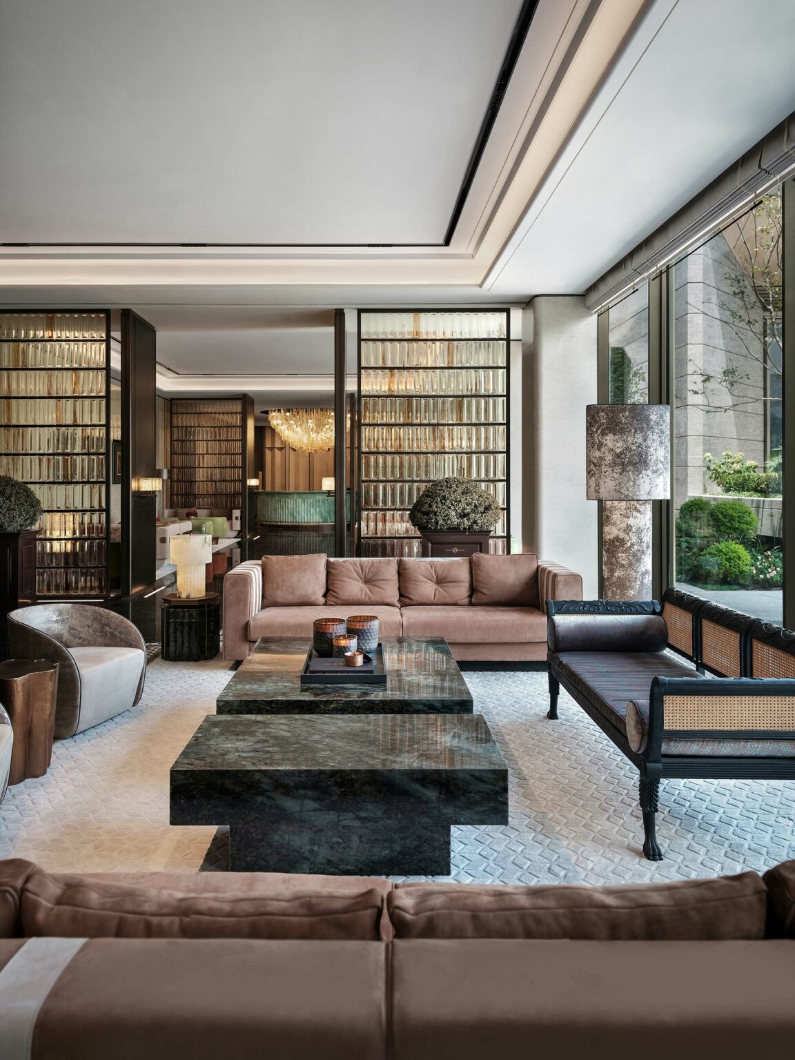 Elegant living room with plush beige sofas, marble tables, and a large window. Shelves full of books and a chandelier add sophistication and warmth.