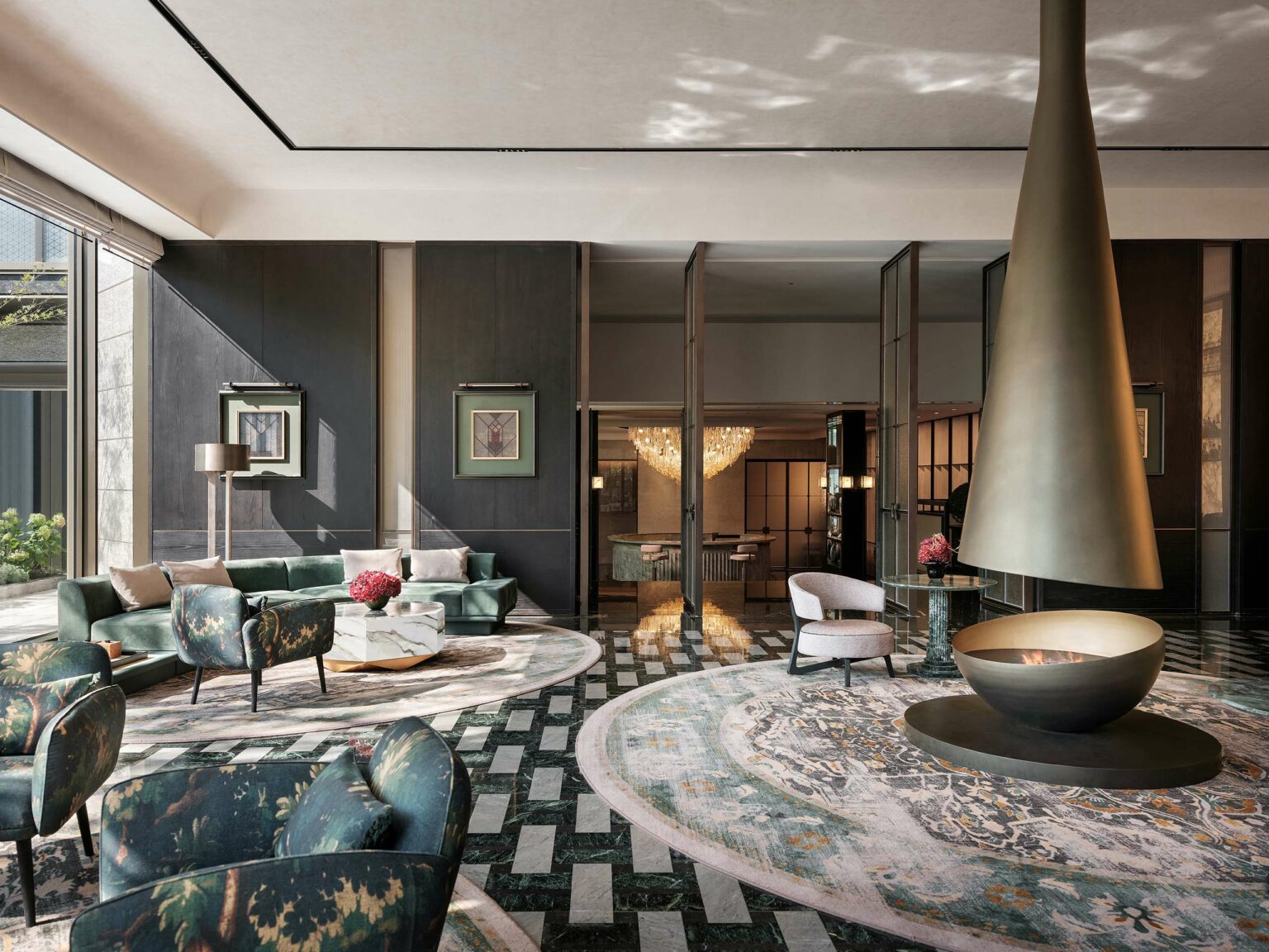 Elegant modern lounge with green armchairs, floral rug, and central conical fireplace. Soft lighting, chic decor, and a luxurious chandelier enhance the ambiance.