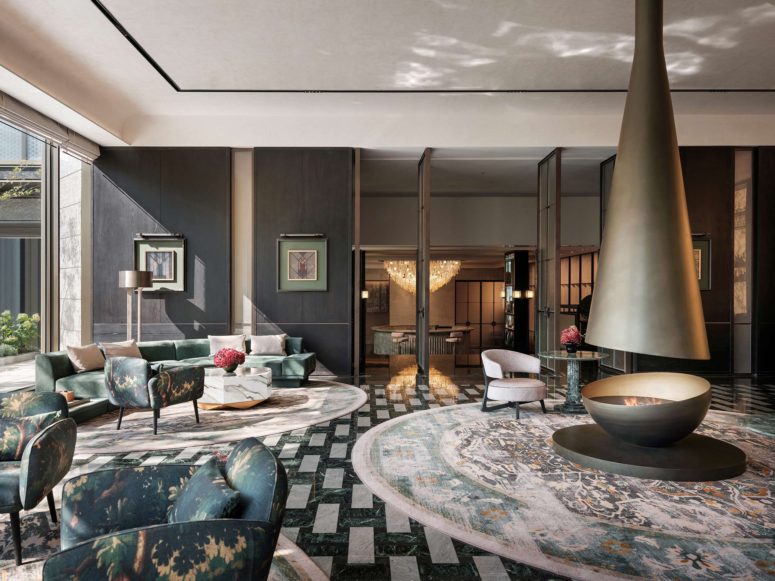 Elegant modern lounge with green armchairs, floral rug, and central conical fireplace. Soft lighting, chic decor, and a luxurious chandelier enhance the ambiance.