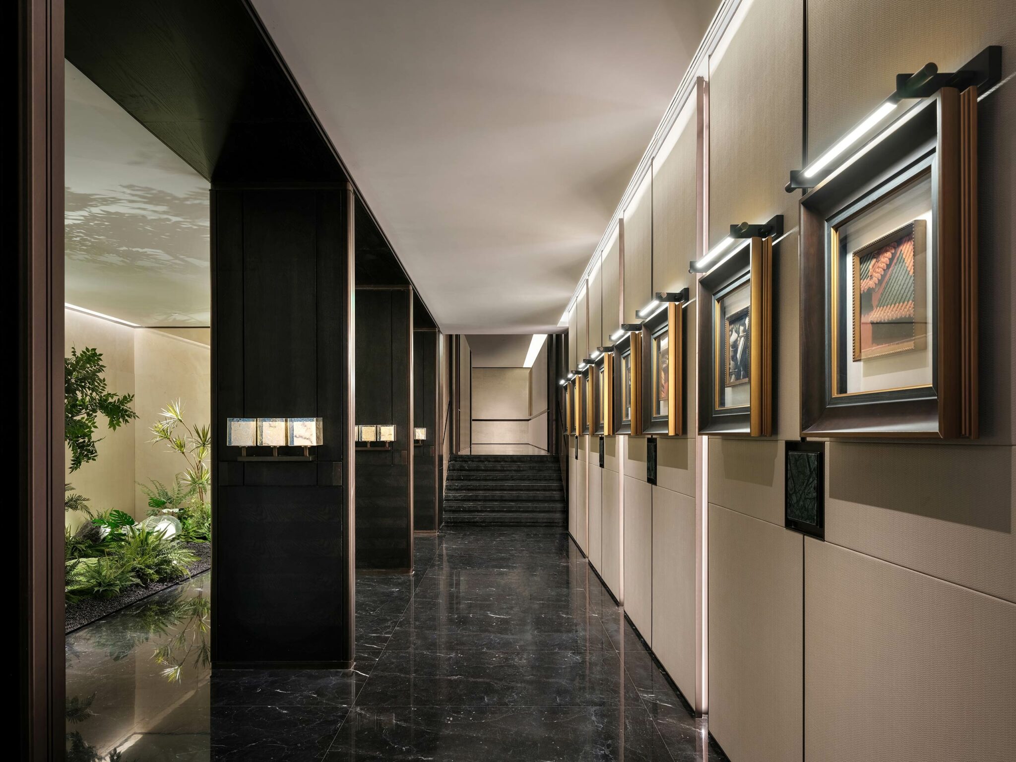 Luxurious hallway with dark marble floor, illuminated framed art on beige walls, and plants in a corner. A staircase leads upward, creating an elegant ambiance.