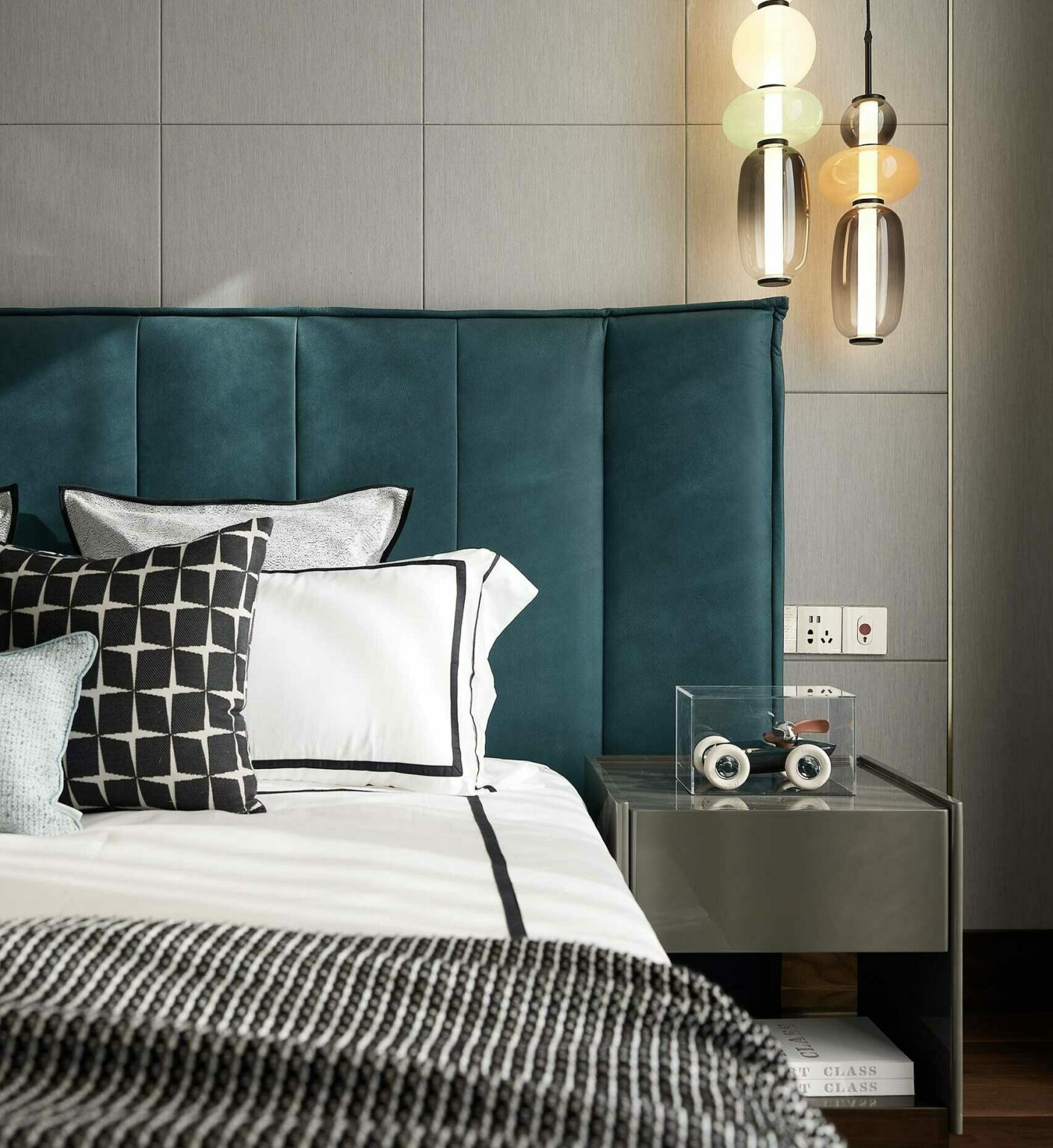 A modern bedroom features a teal headboard, monochrome patterned pillows, and layered bedding. Warm pendant lights hang above a sleek nightstand.