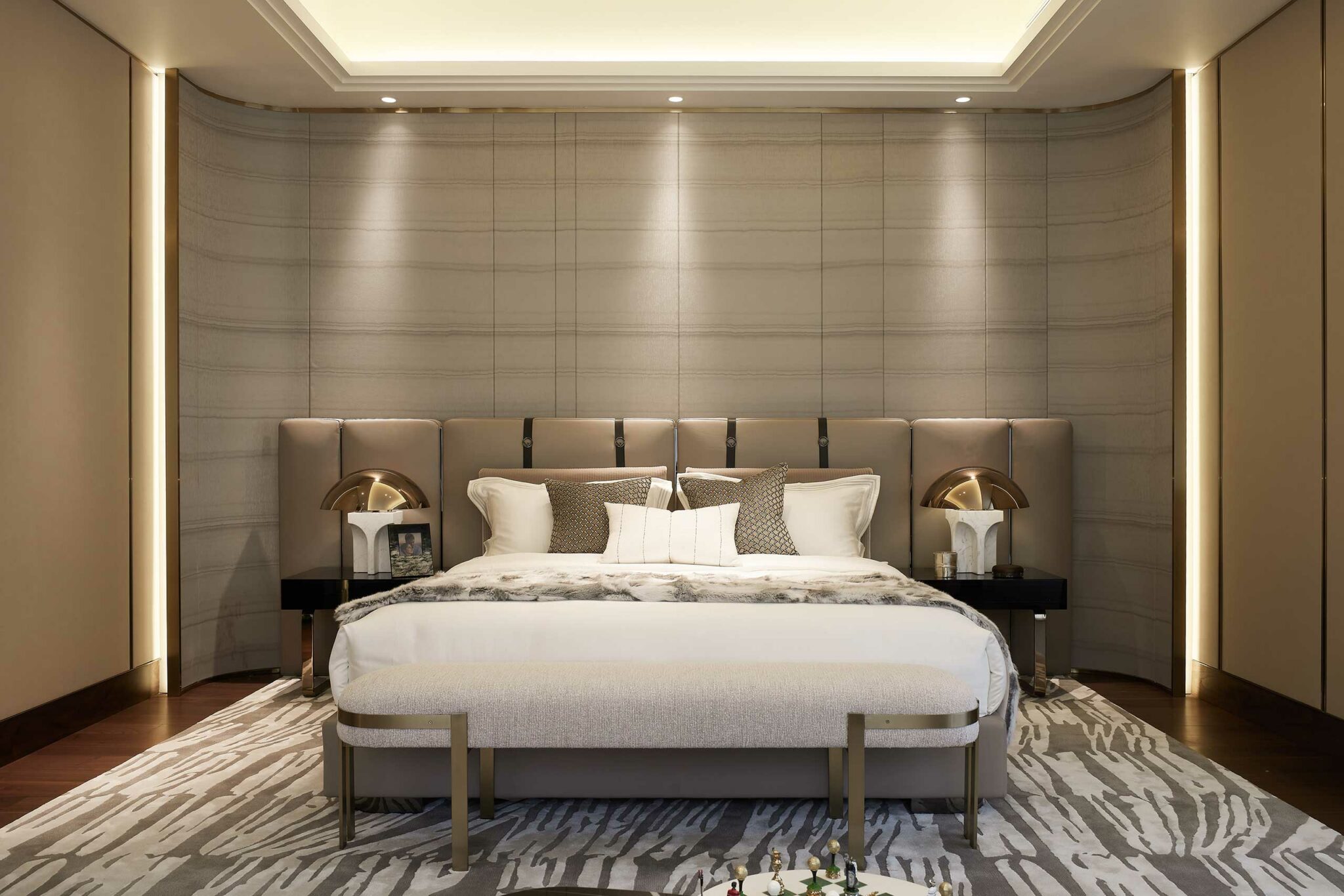Luxurious bedroom with a modern king-size bed, decorative pillows, soft beige tones, elegant lamps on side tables, and a patterned rug, conveying comfort.