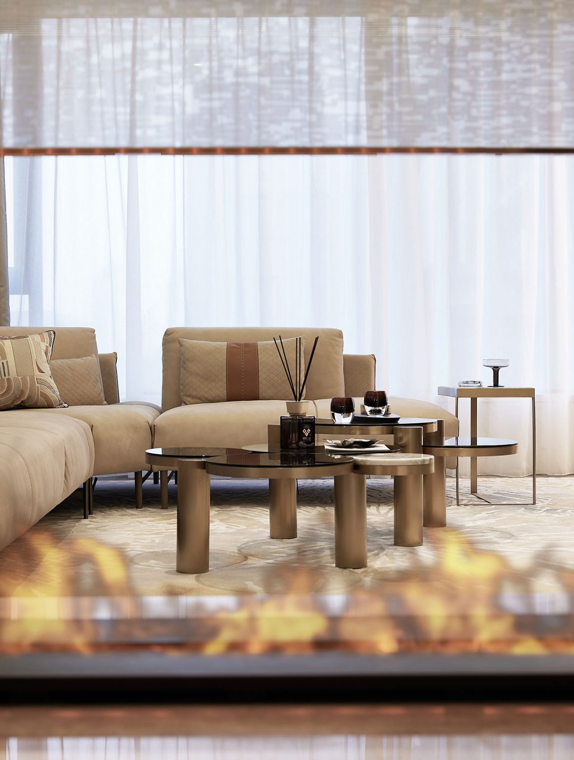 Stylish living room with beige sofas and a glass coffee table, viewed through a glass fireplace. Soft lighting and sheer curtains evoke a cozy atmosphere.