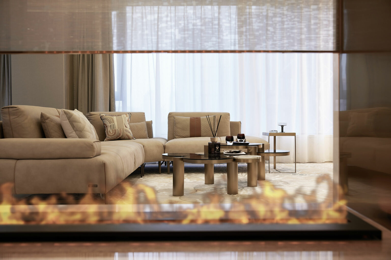 Stylish living room with beige sofas and a glass coffee table, viewed through a glass fireplace. Soft lighting and sheer curtains evoke a cozy atmosphere.