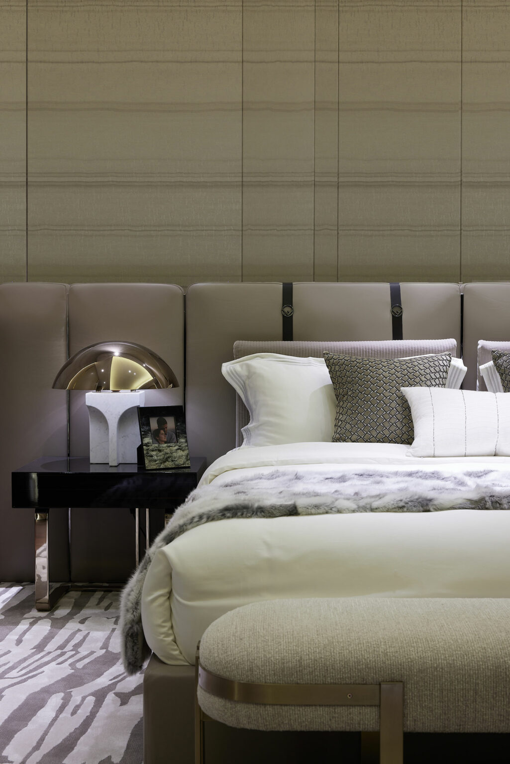 Elegant bedroom with neutral tones. Features a luxurious bed with white and patterned pillows, fur throw, beside a sleek table lamp and a framed photo.