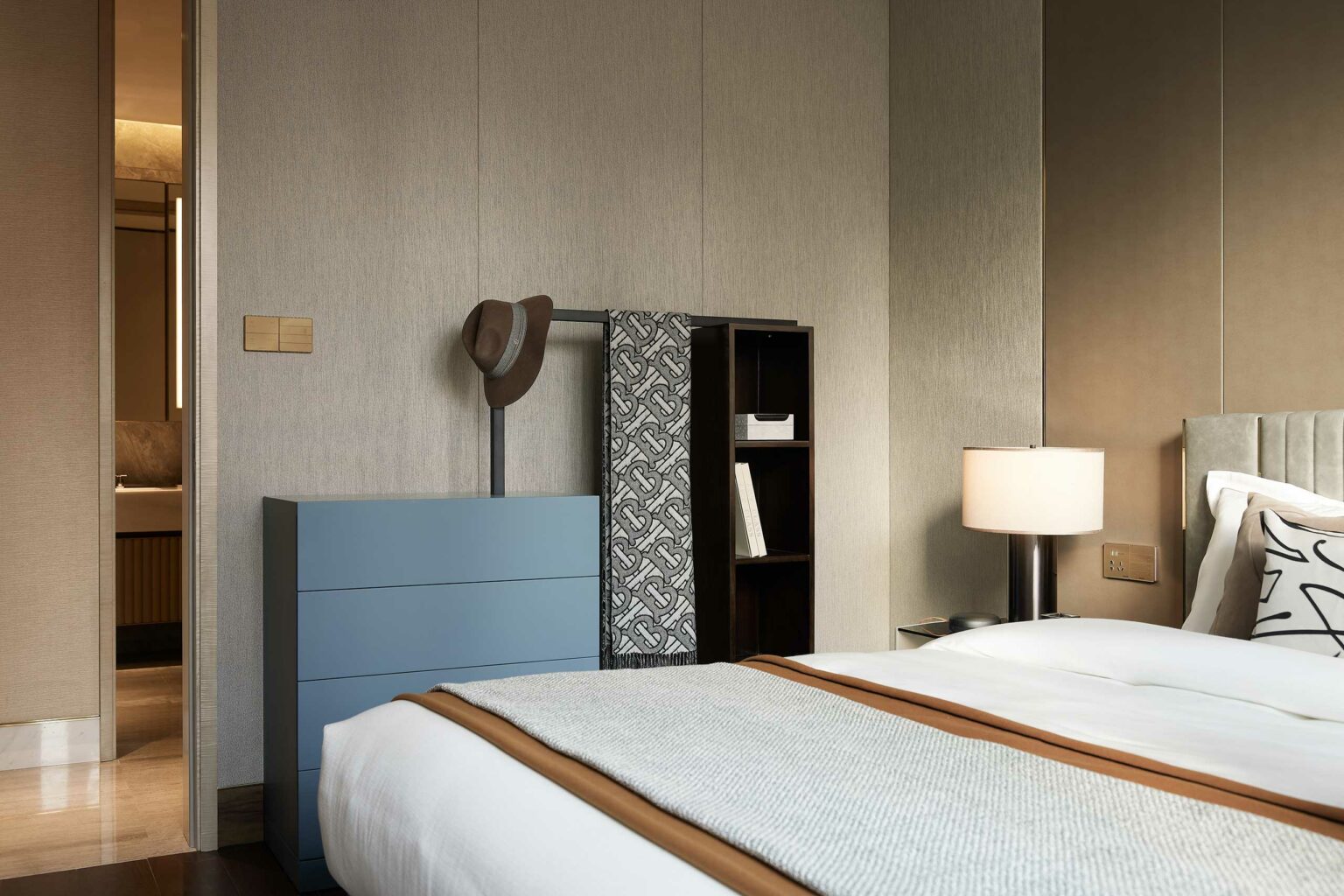 Modern bedroom with a neatly made bed, beige and soft gray tones, a blue dresser with a hat, patterned scarf, and a warm table lamp. Cozy ambiance.
