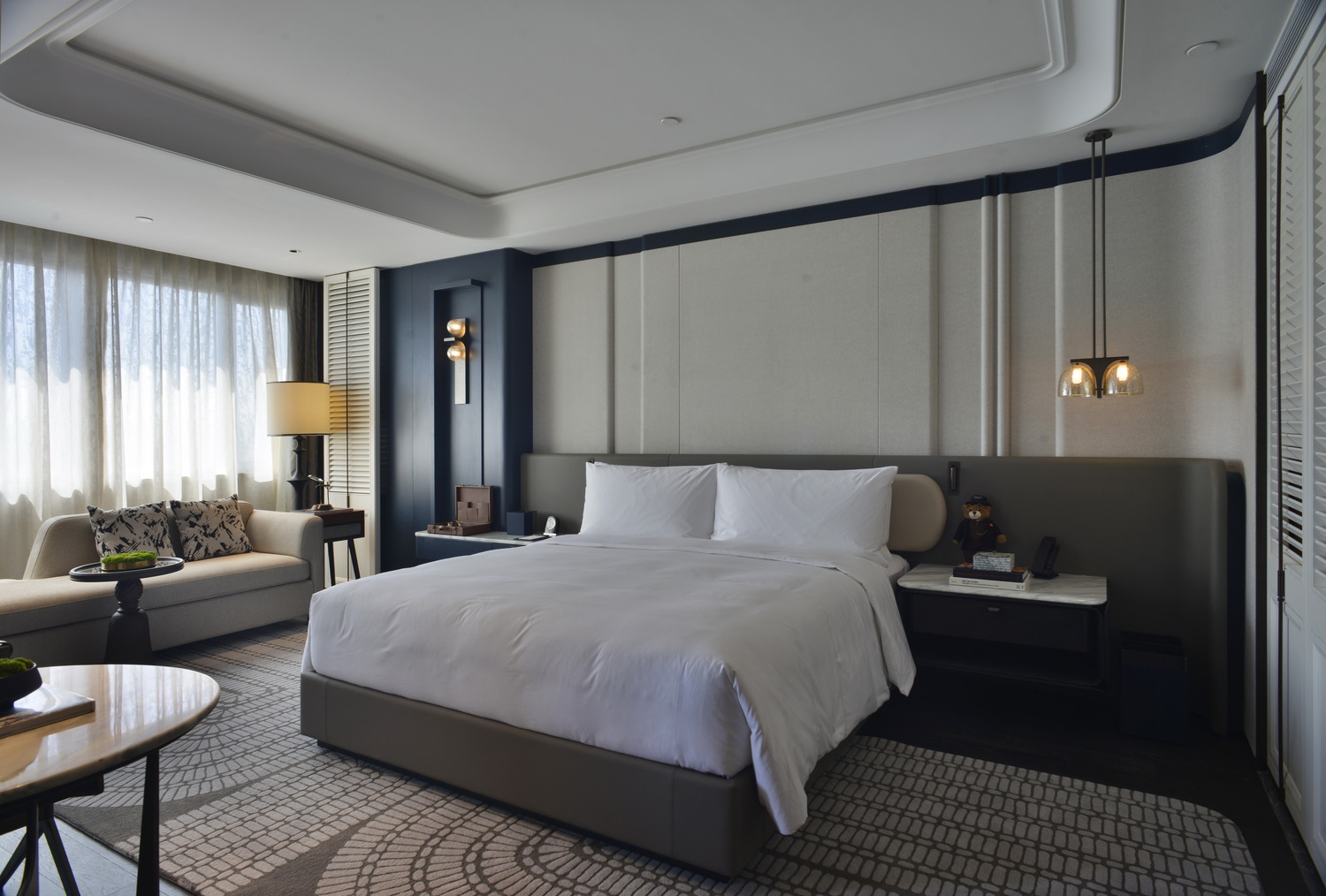 Modern hotel room with two beds, sleek gray tones, and elegant lighting. Features include a seating area, TV, and minibar, creating a cozy yet sophisticated ambiance.