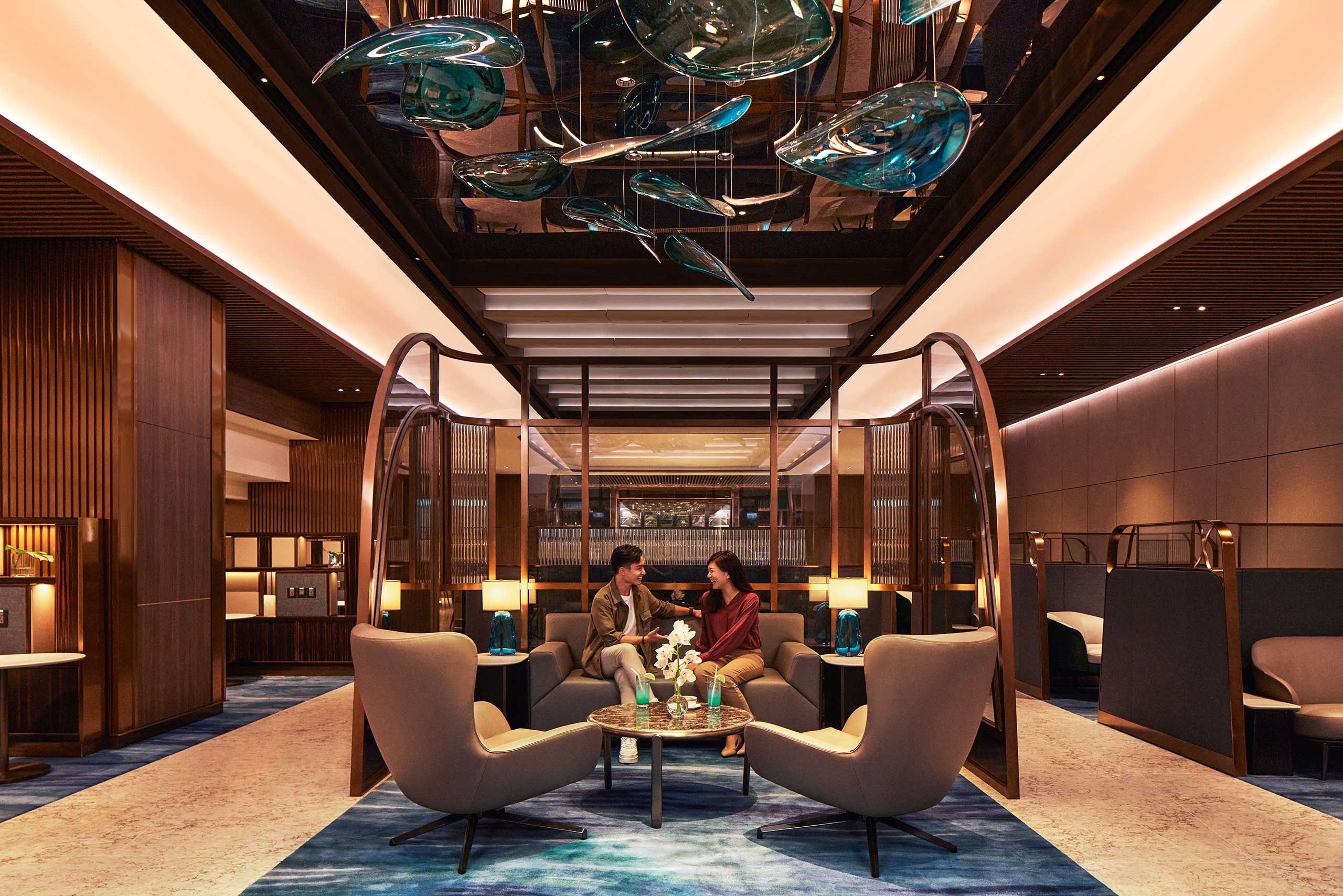 Luxurious airline lounge with modern decor and warm lighting. A couple sits on a sofa, engaged in conversation. Sculptural ceiling art adds elegance. Airy, calm ambiance.