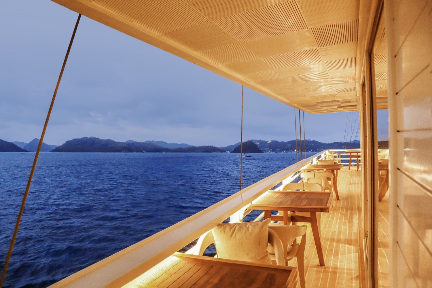 Luxurious yacht deck with plush seating and tables arranged along a white cabin. A person stands gazing at serene water and lush green hills.