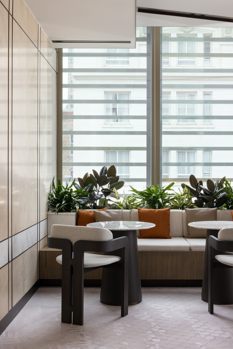 Sophisticated modern office lounge with beige decor, plush seating, and a sleek glass fireplace. Floor-to-ceiling windows and greenery create an airy, relaxing atmosphere.