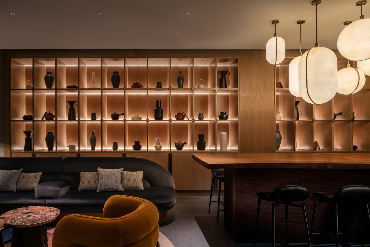 Modern hotel lounge with warm lighting, featuring a backlit wooden display of vases, a curved sofa with pillows, mustard armchair, wooden bar, and pendant lights.