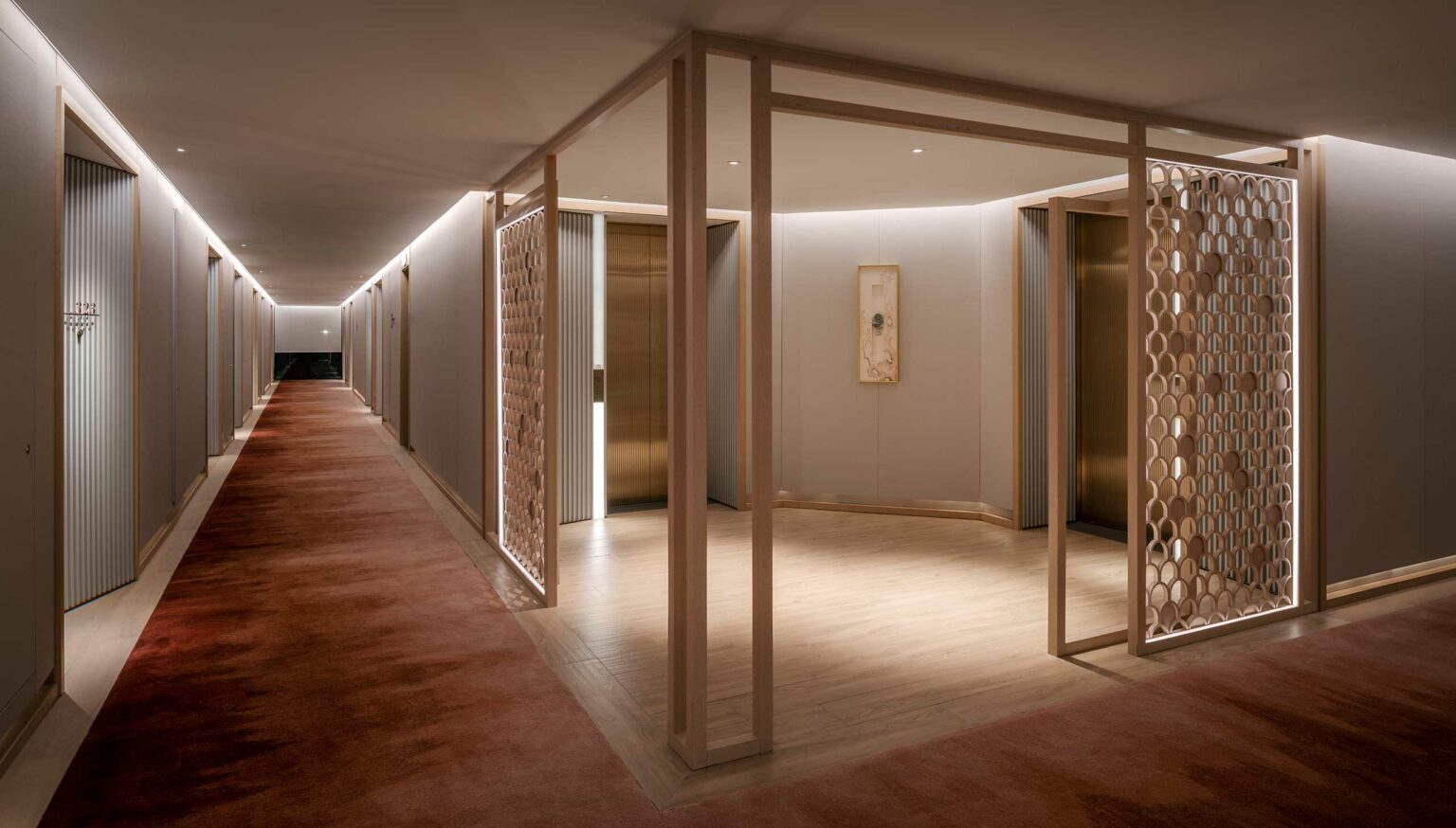A modern, elegantly-lit hotel hallway features a warm, red carpet. Wooden lattice panels and vertical lines accentuate the decor, creating a serene and inviting ambiance.