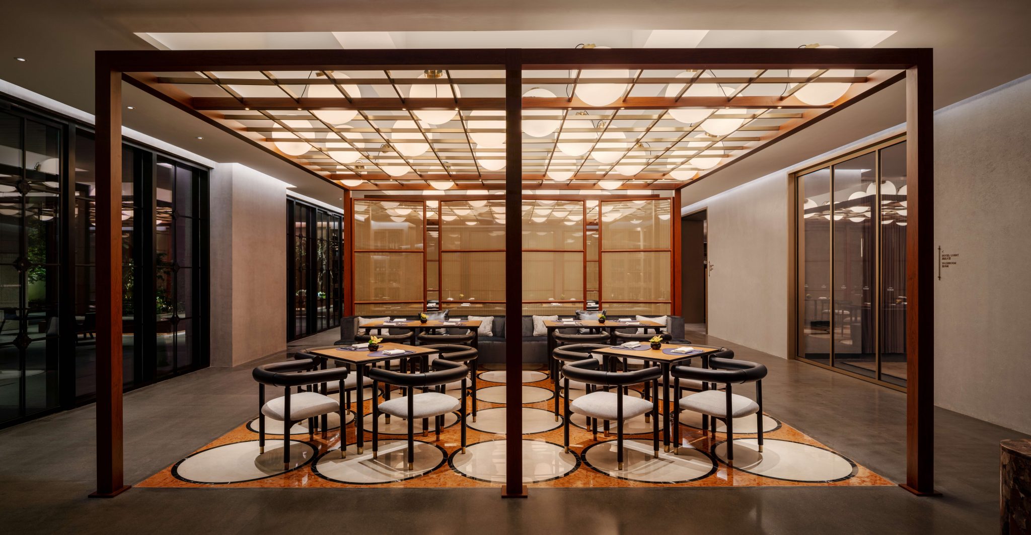 Modern hotel lounge with warm lighting, featuring a backlit wooden display of vases, a curved sofa with pillows, mustard armchair, wooden bar, and pendant lights.
