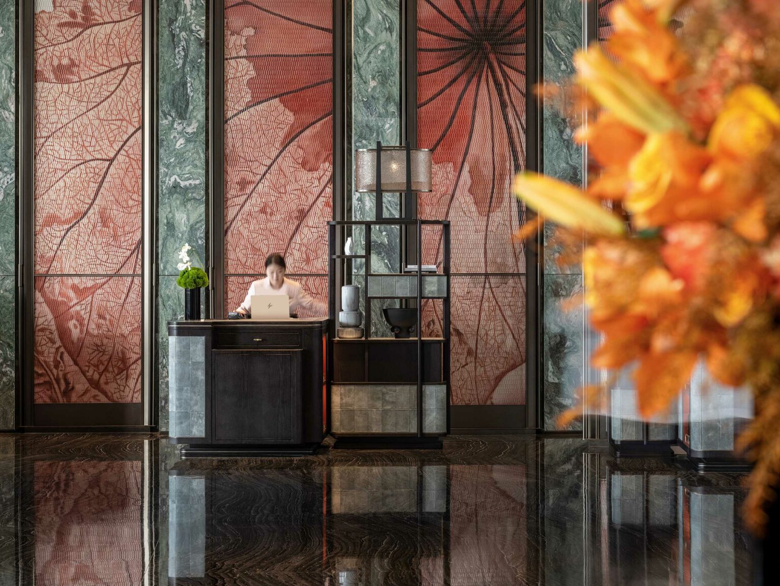 A hotel reception area with a person at a desk using a laptop. The background has elegant red and green leaf-patterned panels. Bright orange flowers are in the foreground.