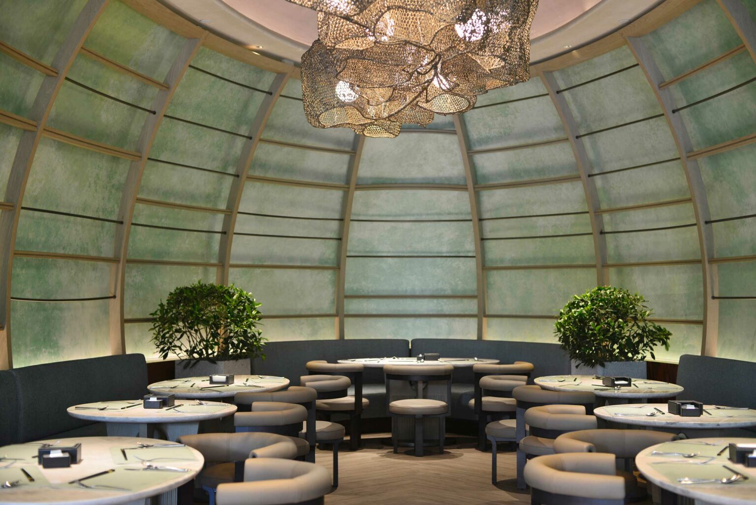 Modern hotel restaurant interior with a dome-shaped ceiling and a large, artistic chandelier. Round tables, cushioned stools, and greenery create an inviting feel.