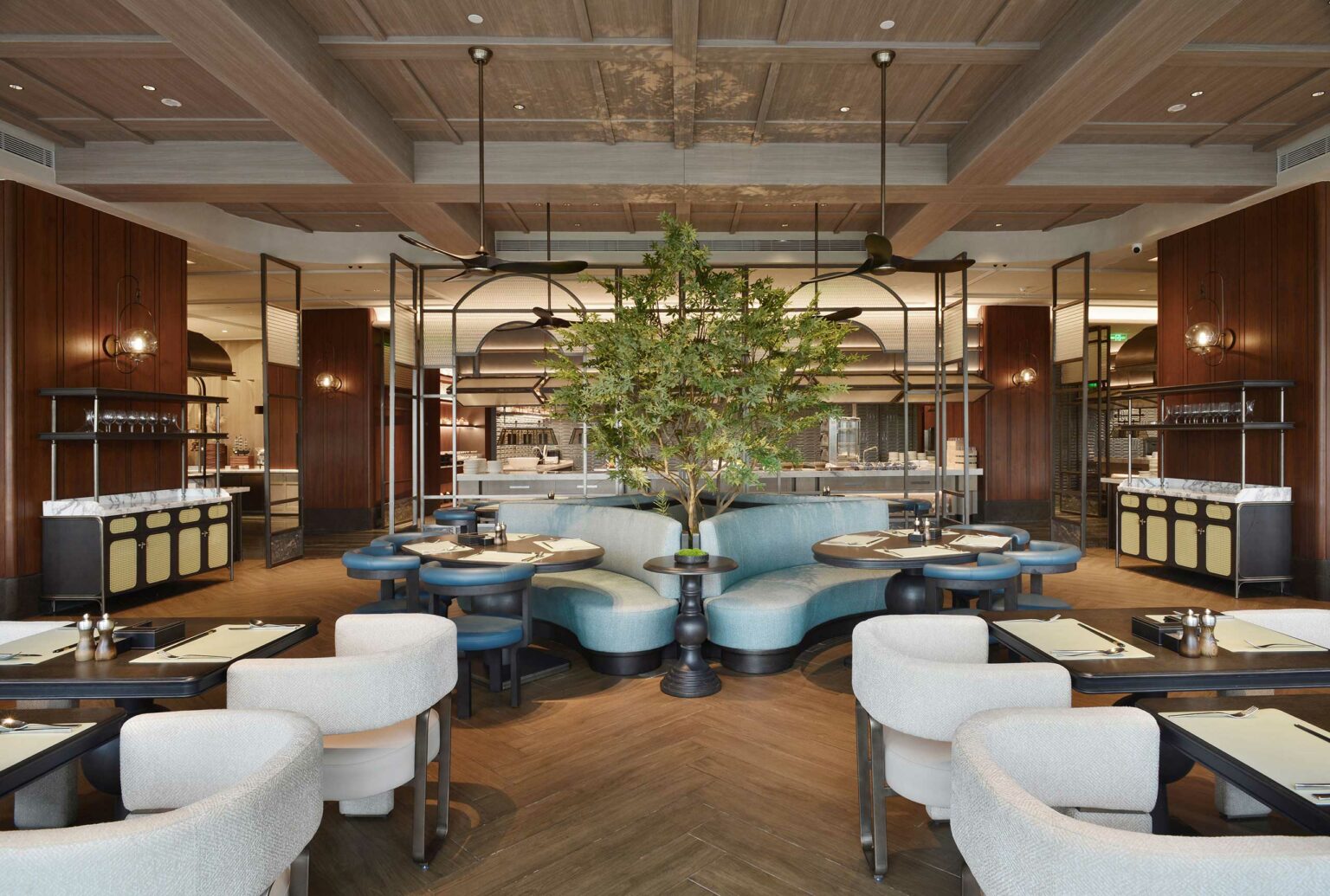 Modern hotel dining area with a central tree, curved blue booths, and white chairs on a wooden floor. Warm lighting creates an inviting atmosphere.