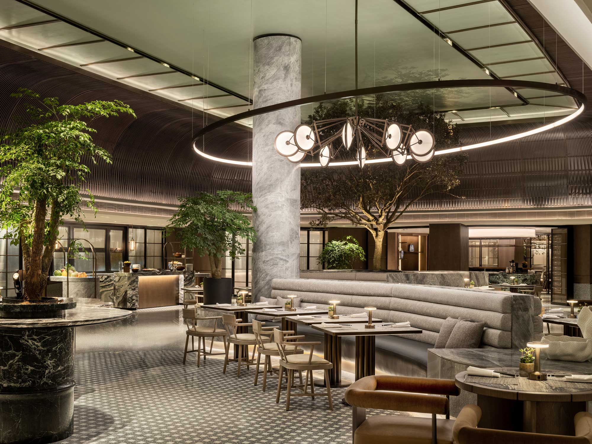 Sophisticated hotel lobby with modern decor featuring lush indoor trees, circular chandelier, marble columns, and curved seating. Cozy and elegant ambiance.