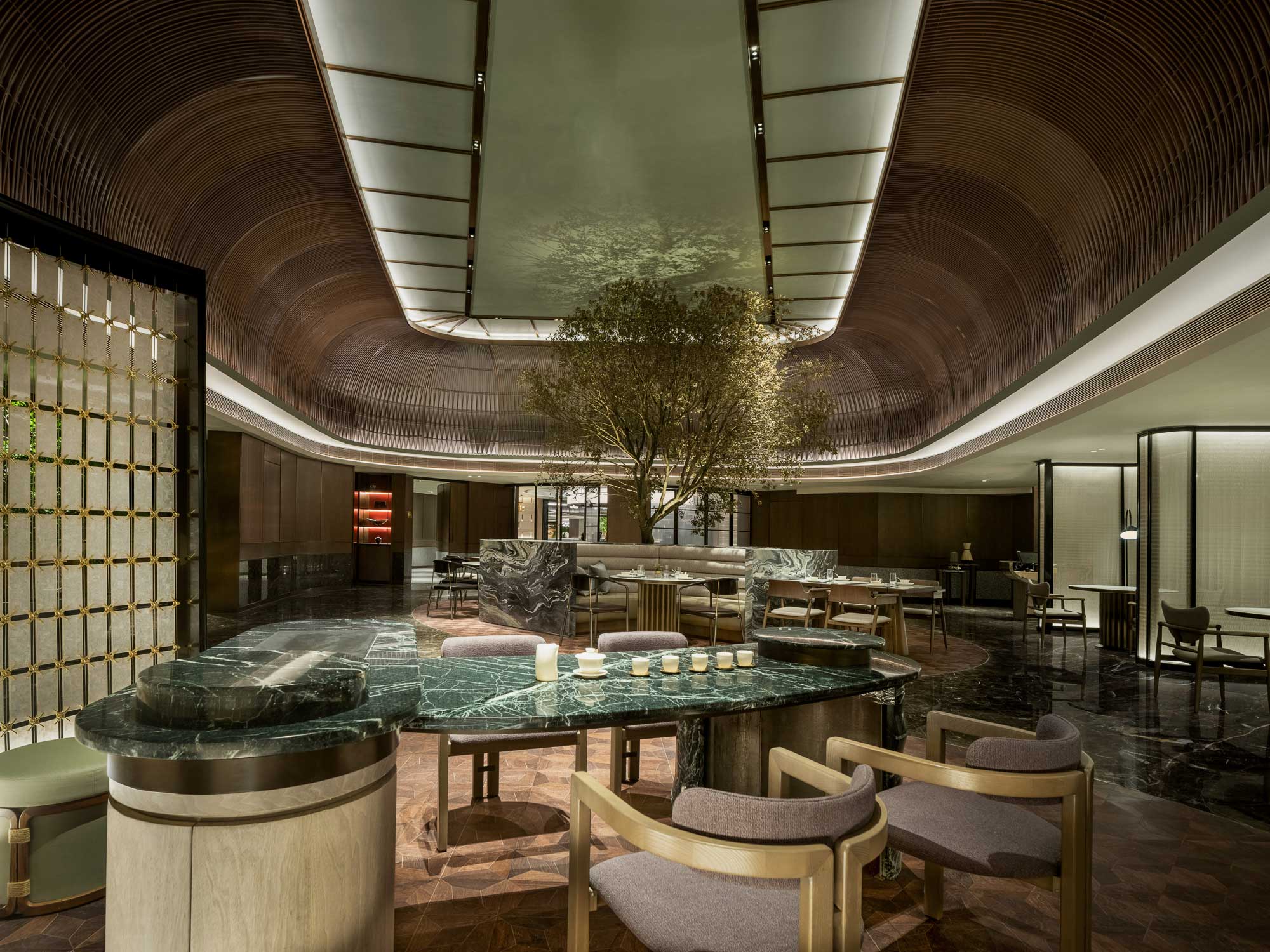 Luxurious hotel lobby with modern decor, featuring a central tree, marble tables, plush chairs, warm lighting, and a curved, wood-paneled ceiling.