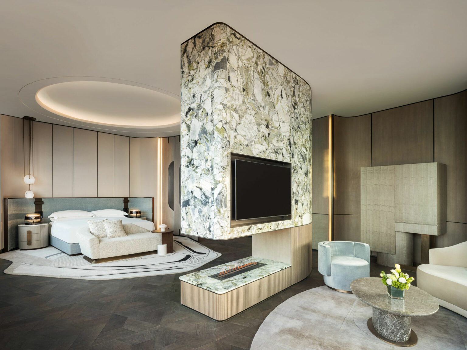Luxurious hotel suite with a modern design. A marble room divider holds a flat-screen TV and separates the bed from the lounge area with plush seating and a round table.