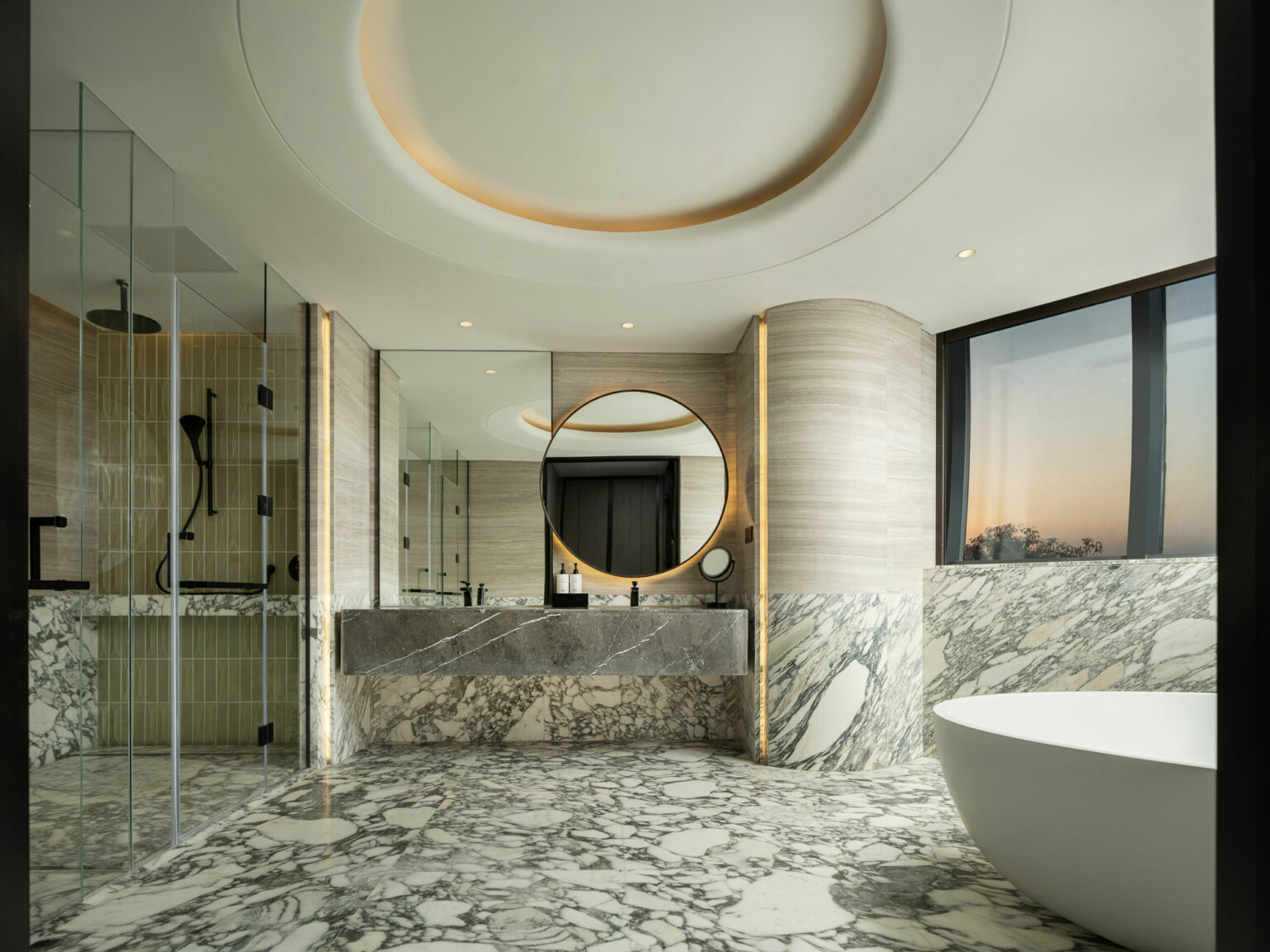 Luxurious suite bathroom with circular mirror, marble walls, and floor. Spacious shower, freestanding tub, and large windows showing a serene sunset.