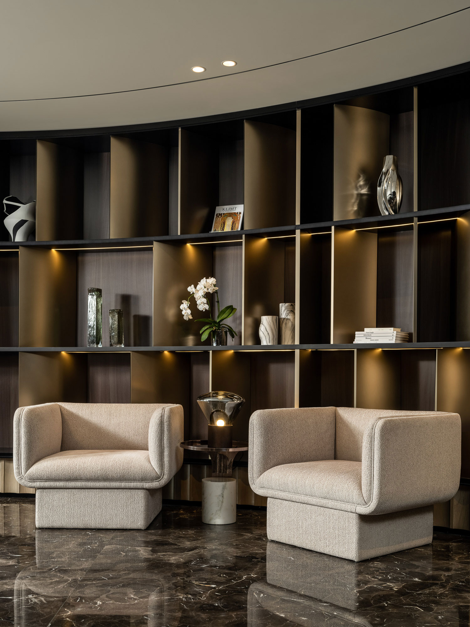 Modern hotel lounge with two beige armchairs on a dark marble floor. Behind, a curved wooden shelf displays vases, plants, and books, creating a serene ambiance.