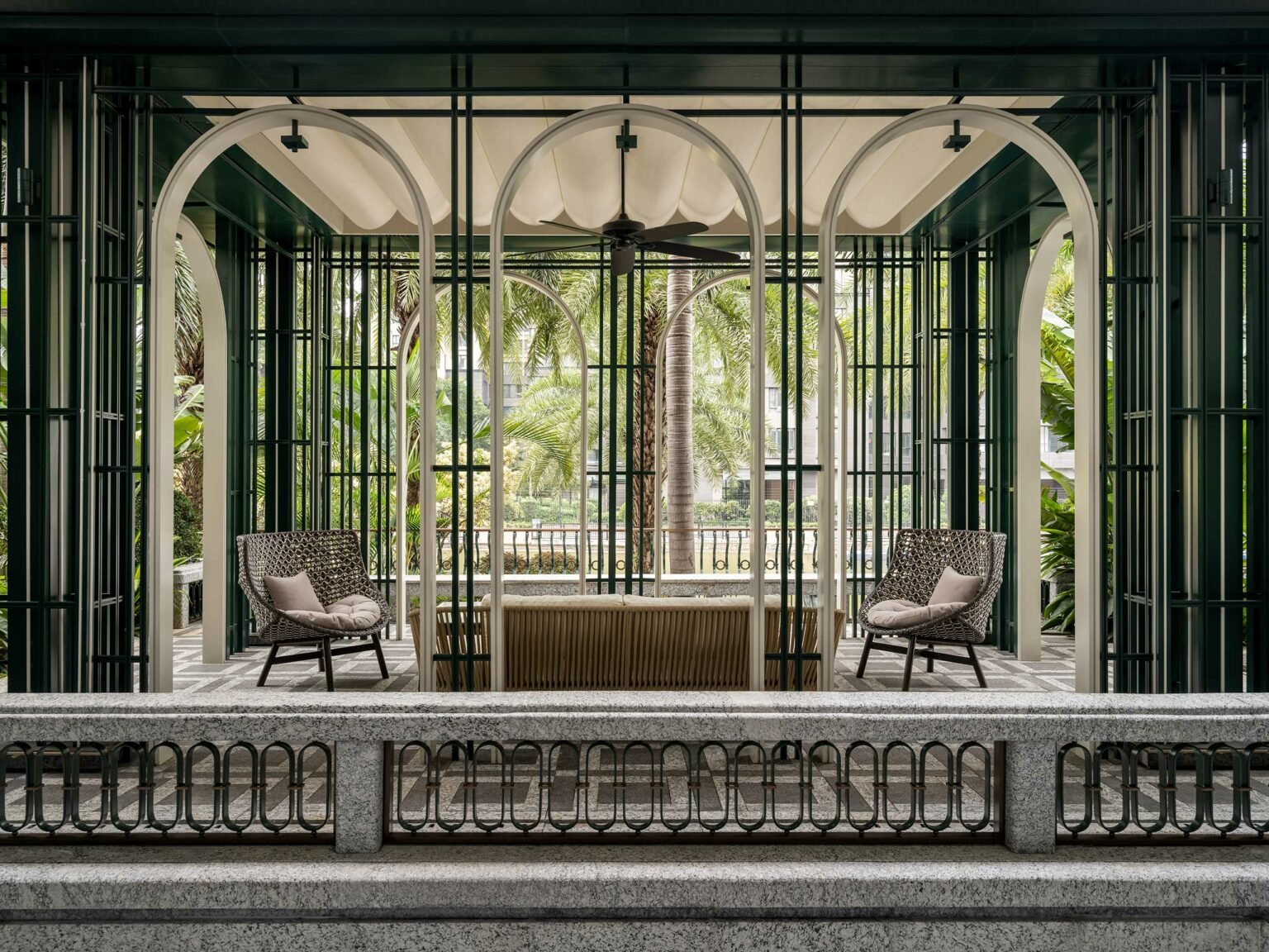 Elegant hotel outdoor space with two patterned chairs and a table under an arched pavilion. Tropical plants and palm trees create a serene atmosphere.