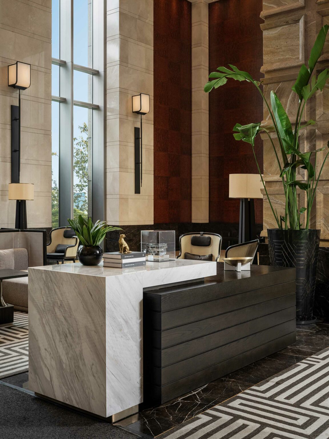 Elegant hotel lobby with modern decor. Features a marble desk, tall plants, large windows, neutral tones, and soft lighting, creating a luxurious ambiance.