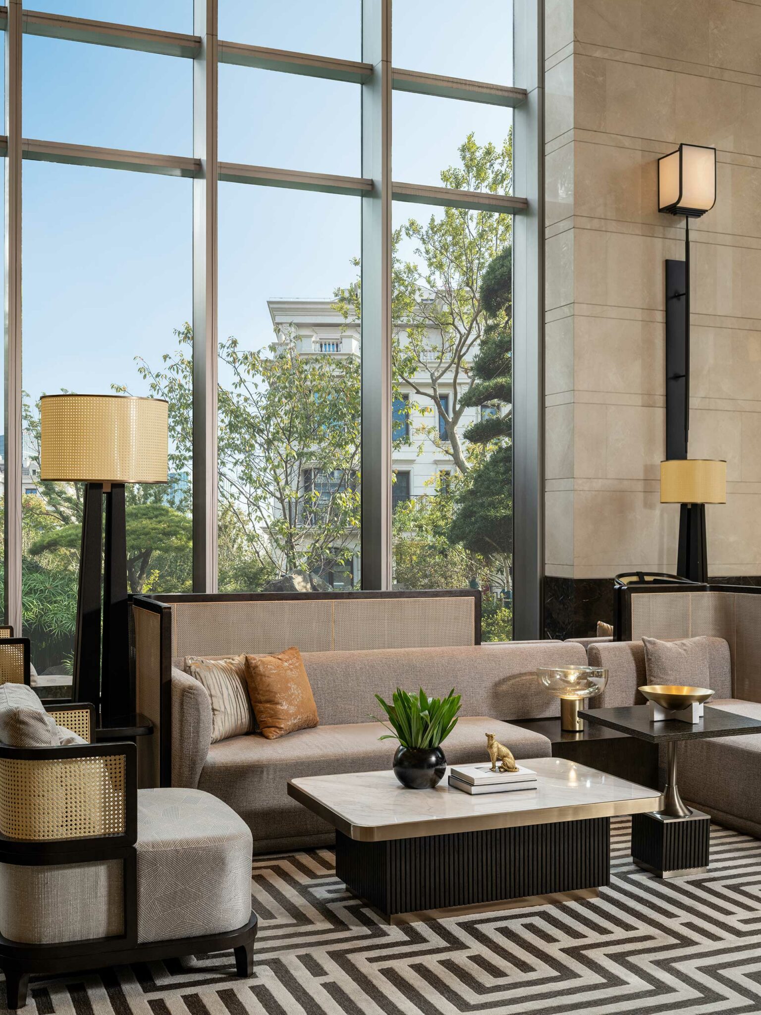 Sophisticated hotel lobby with large windows, beige sofas, and modern lamps. A marble coffee table features a green plant, books, and decorative items.