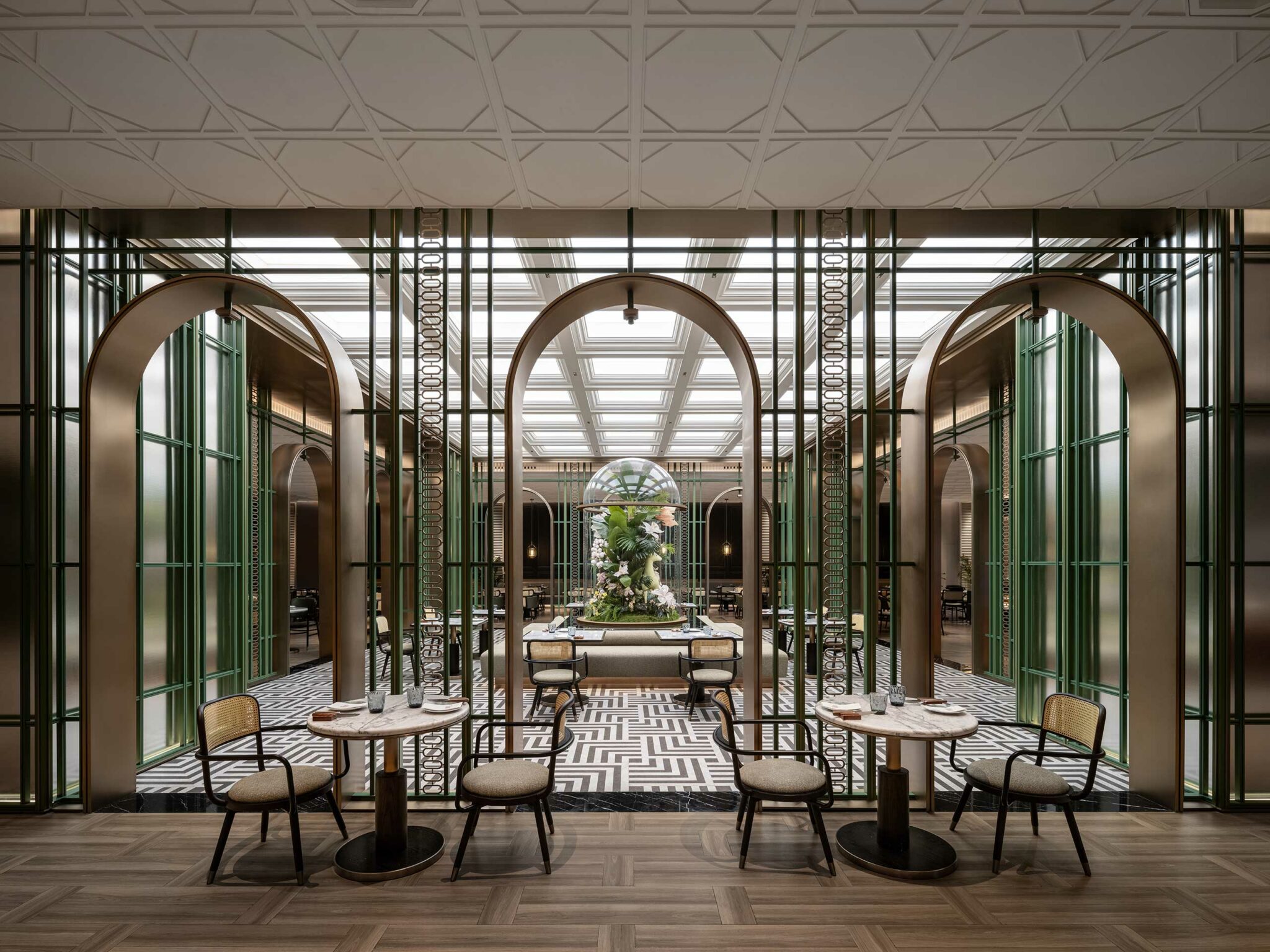 Luxurious hotel restaurant interior with geometric tile flooring, wooden chairs, and tables. Central archways lead to a floral sculpture under glass. Sophisticated ambiance.