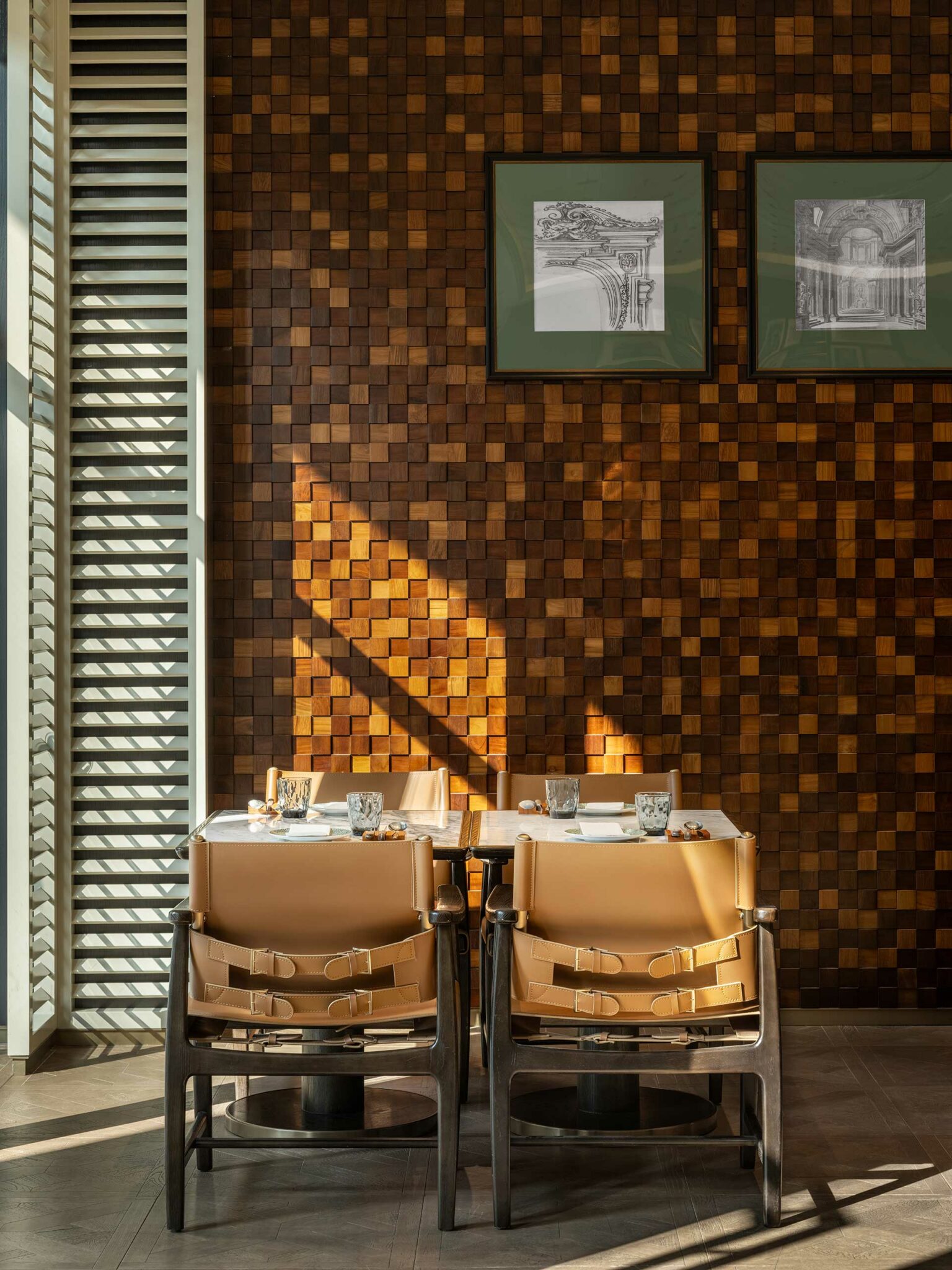 Sunlight casts geometric patterns on a textured wooden wall with framed sketches. Two tan chairs face a set table, creating a warm, inviting tone.