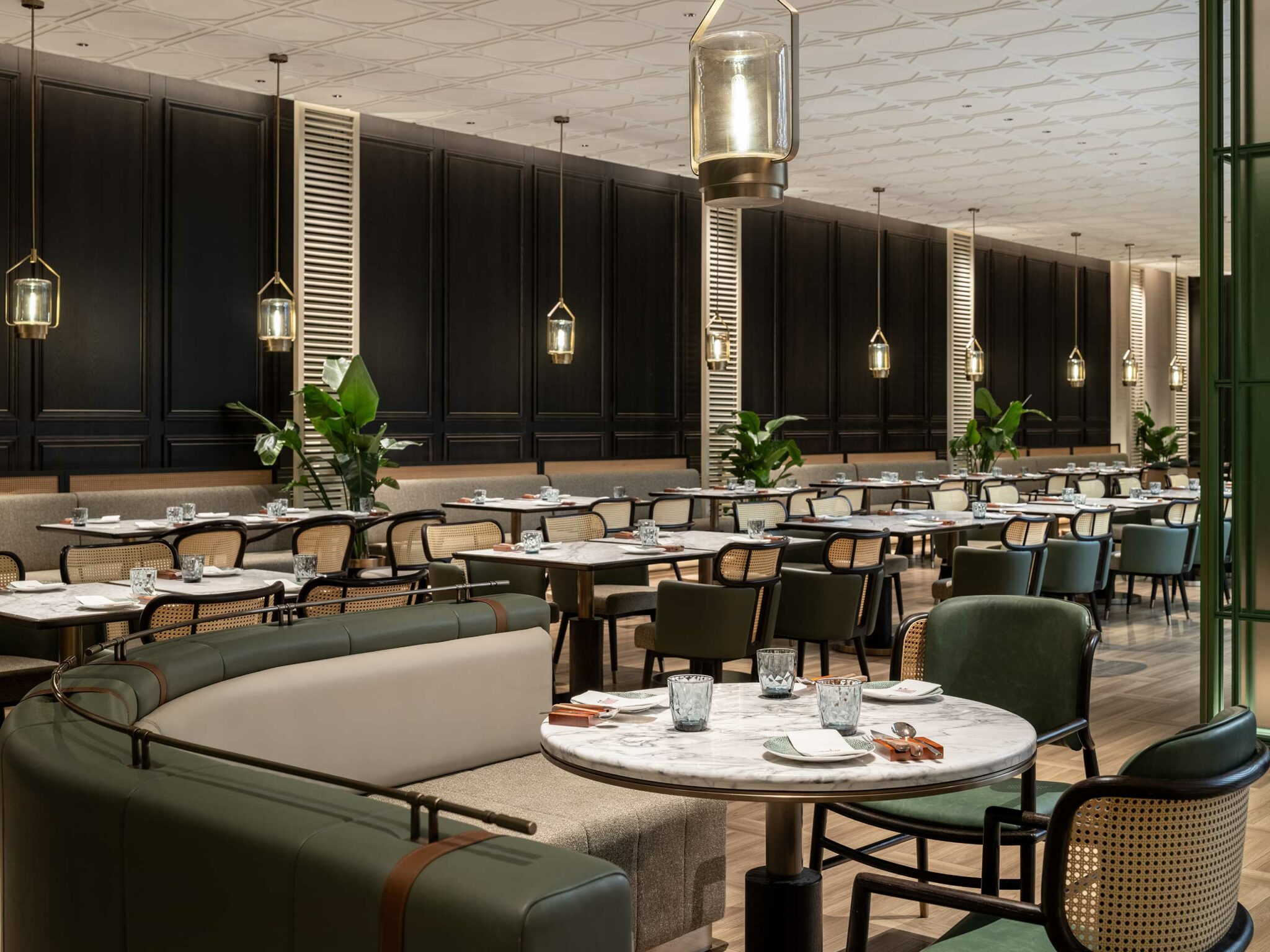 Elegant hotel restaurant interior with marble tables, green chairs, and hanging lanterns. Neutral tones, modern decor, and lush green plants create a calming ambiance.