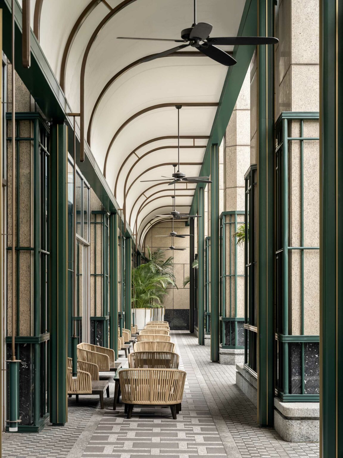Elegant, arched hotel corridor with wicker chairs and ceiling fans, bordered by tall, green-framed windows and lush plants, creating a serene and airy atmosphere.