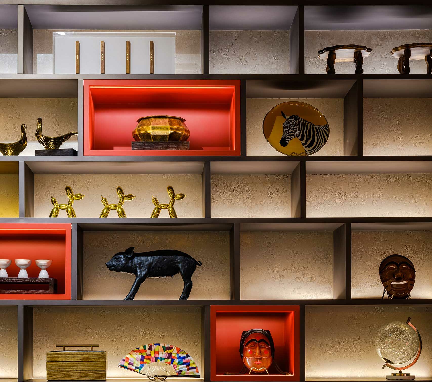 Modern shelf display featuring eclectic decor items like a black pig sculpture, golden duck figurines, and a zebra plate. Red accents add vibrancy.