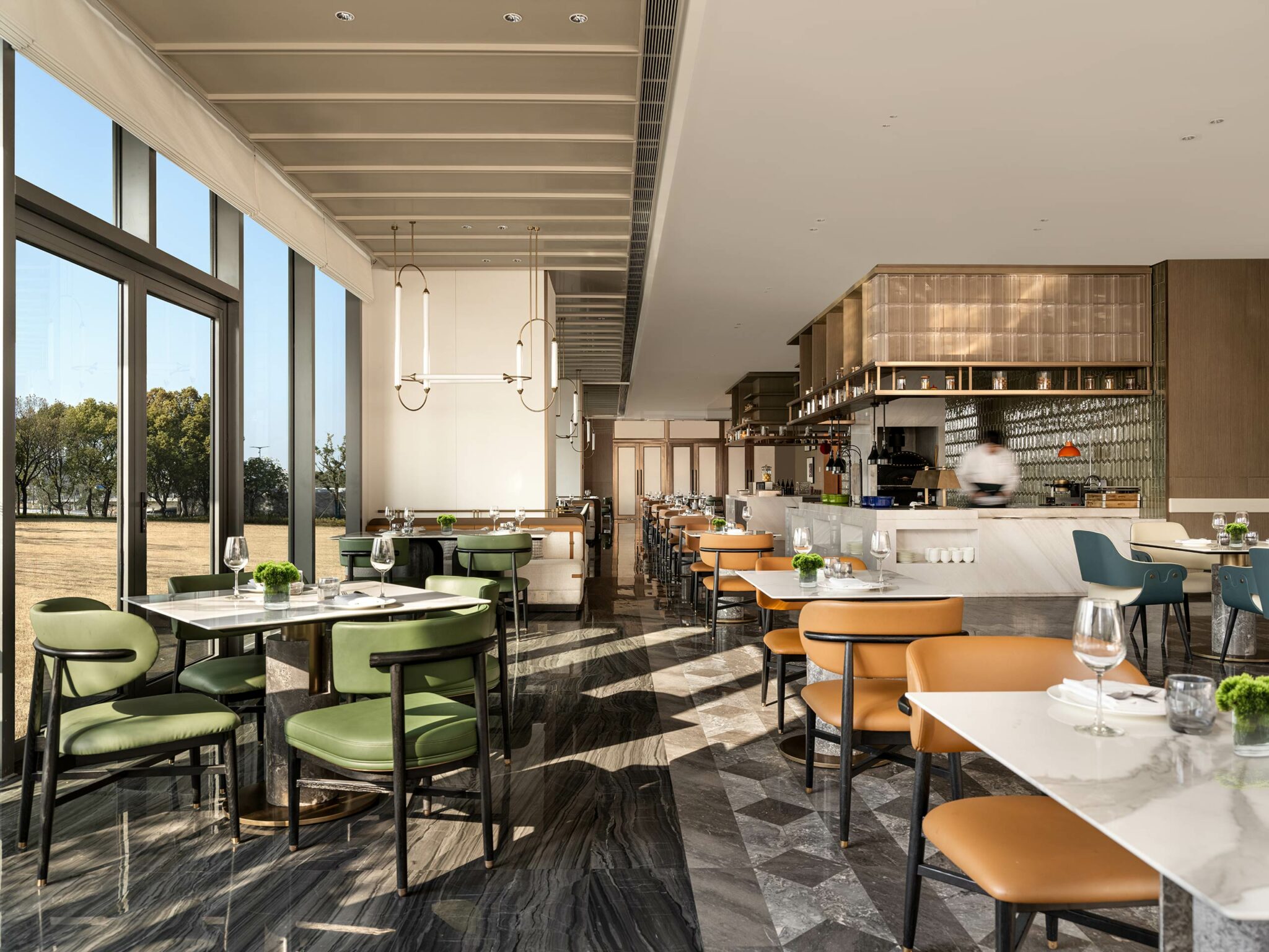 Modern hotel all day dining interior with large windows and abundant natural light. Green and tan chairs surround tables set with wine glasses; open kitchen visible.