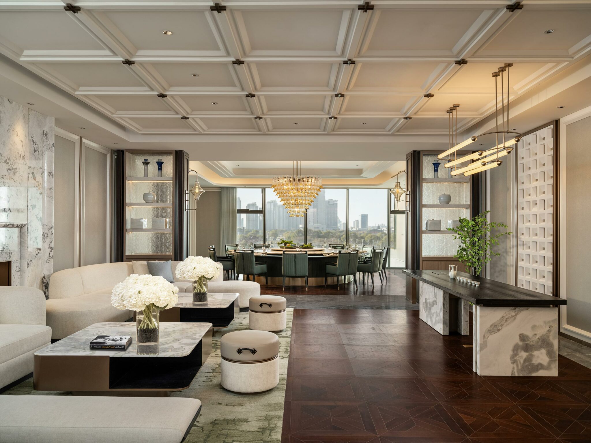 Luxurious open-plan hotel dining area with elegant seating, floral centerpieces, a large chandelier, and a city skyline view through expansive windows.