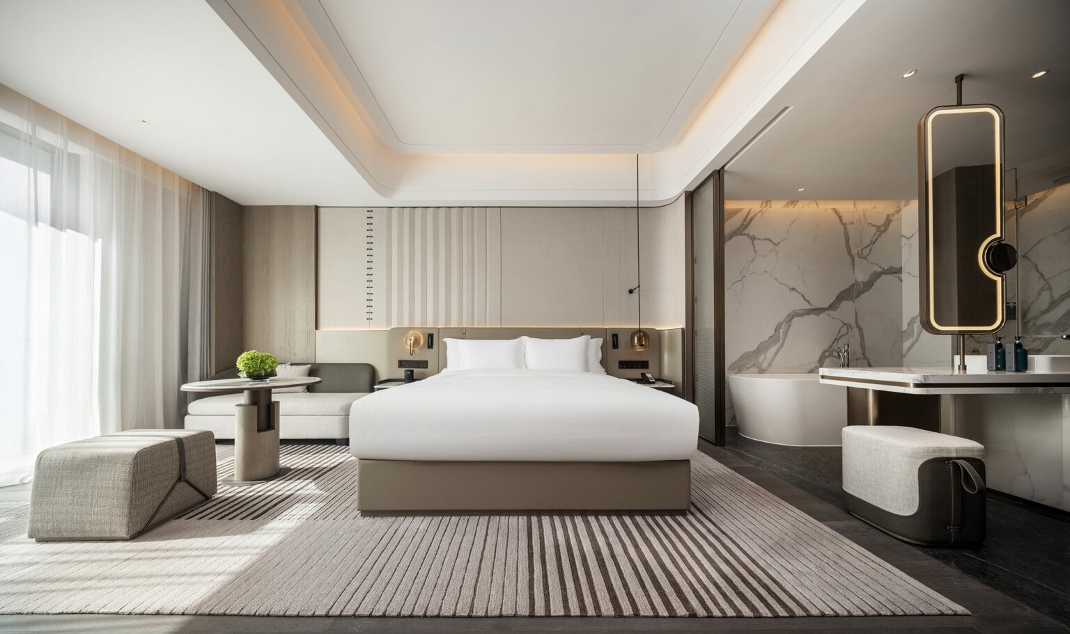 Modern hotel room with neutral tones featuring a large bed, marble bathroom with a freestanding tub, sleek furniture, and soft lighting, conveying relaxation.