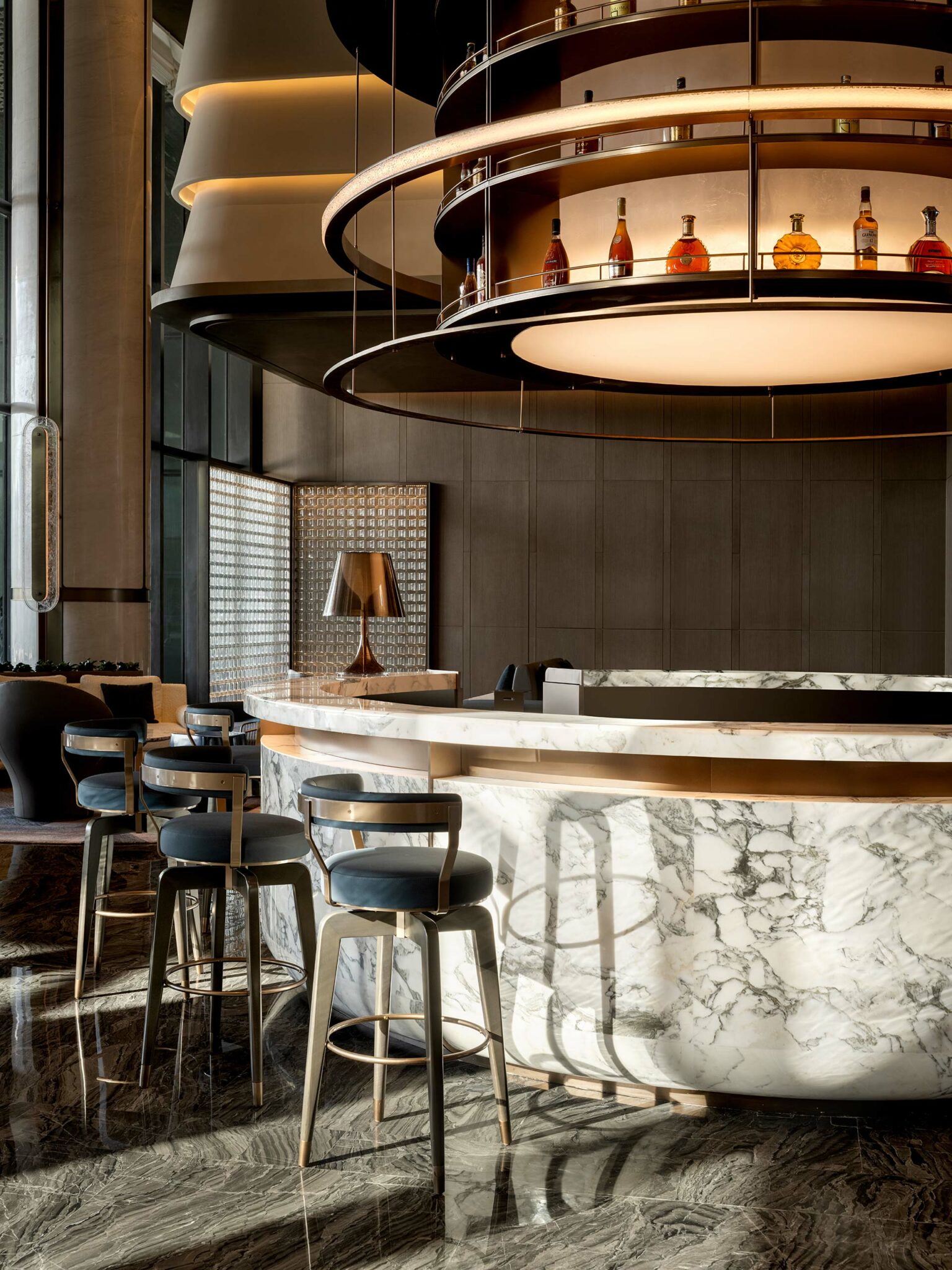 Luxurious hotel bar with a circular marble counter, high-backed chairs, and an overhead rack with liquor bottles. Warm lighting creates an elegant atmosphere.