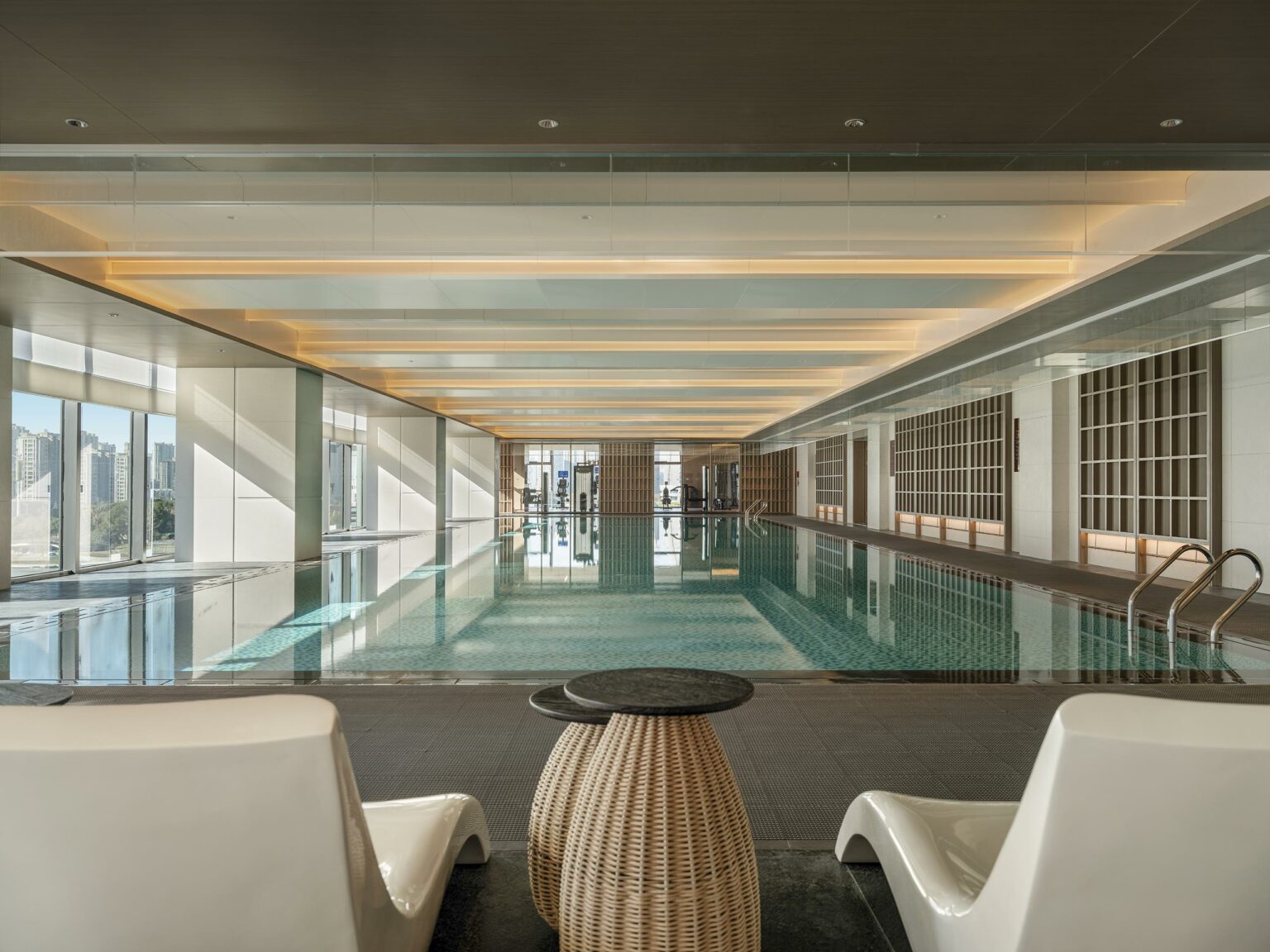 Luxurious hotel indoor pool with calm water, surrounded by white and wooden walls, lit by overhead lights. Lounge chairs and a wicker table are in the foreground.