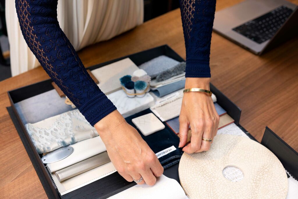 Hands arrange fabric and color samples in a tray on a wooden table, with a laptop nearby. The scene conveys a creative, focused workspace.