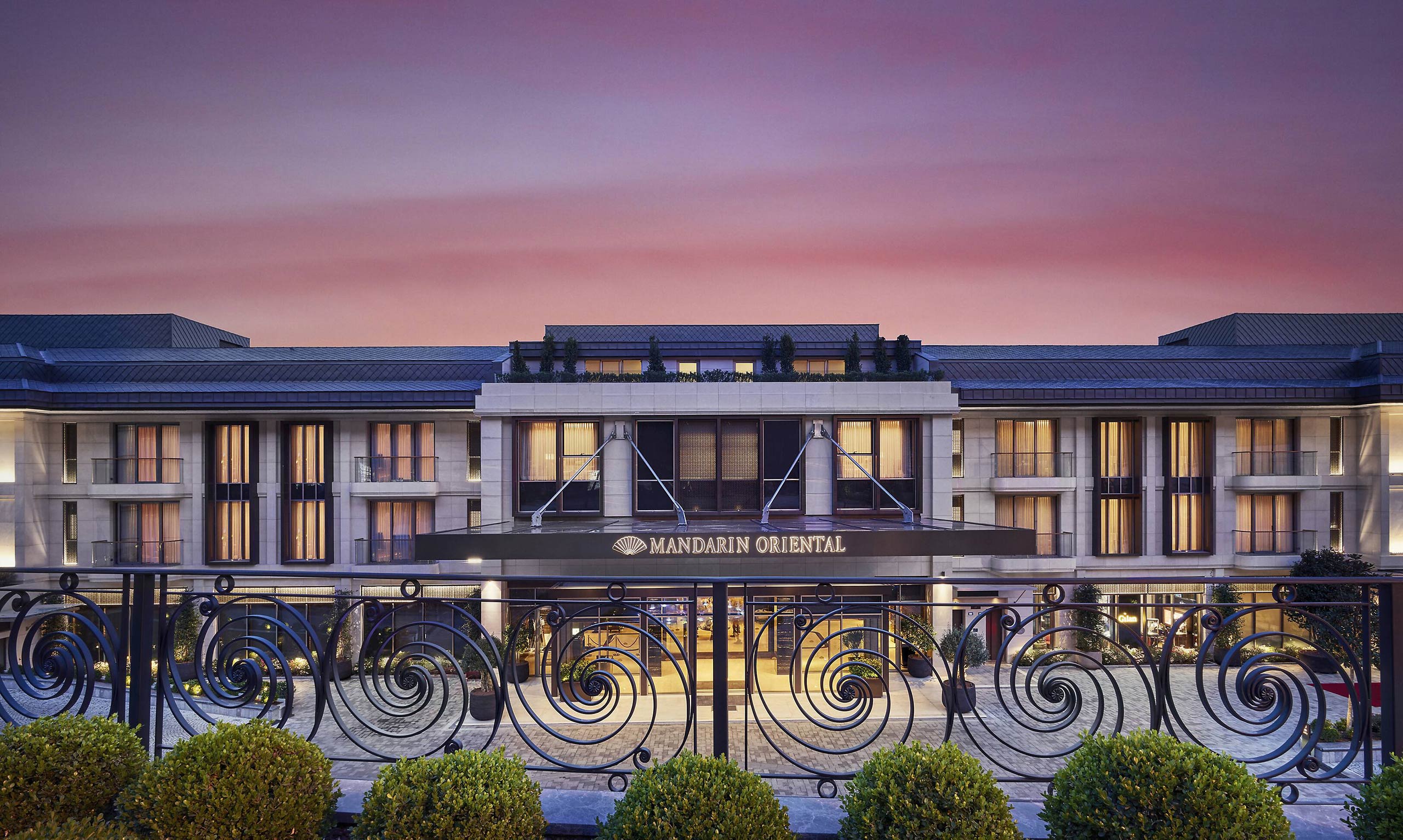 Elegant hotel facade of the Mandarin Oriental Bosphorus Istanbul at dusk, with warm lighting glowing from windows and a soft pink and purple sky, conveying luxury and serenity.
