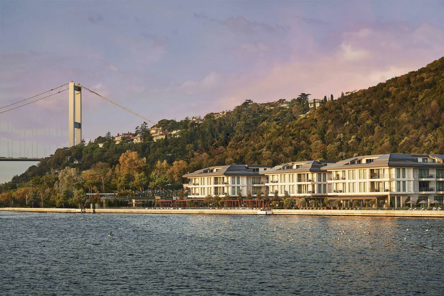 An elegant waterfront hotel in front of a forested hillside and a suspension bridge under a soft pink sky. The scene is tranquil and inviting.