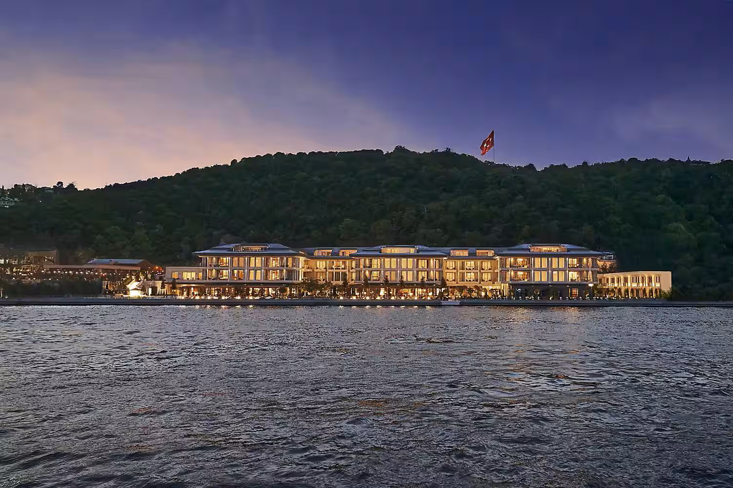 An elegant waterfront hotel is illuminated at dusk against a lush, dark hillside. Its warm lights reflect on the calm water, creating a serene ambiance.