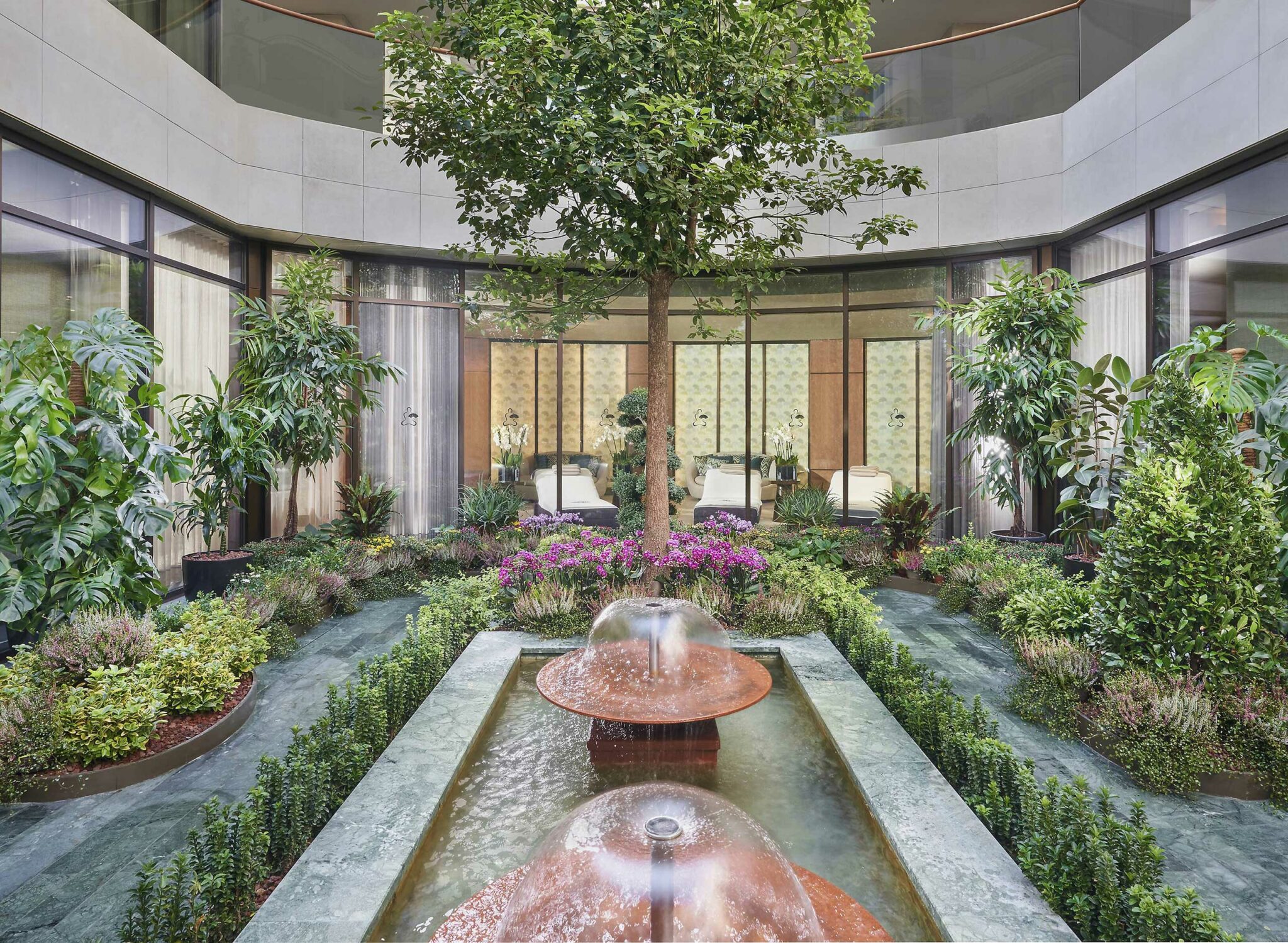 Lush hotel courtyard garden with a central tree and colorful flowers, surrounded by plants. A water feature with fountains adds tranquility, adjacent to glass walls.